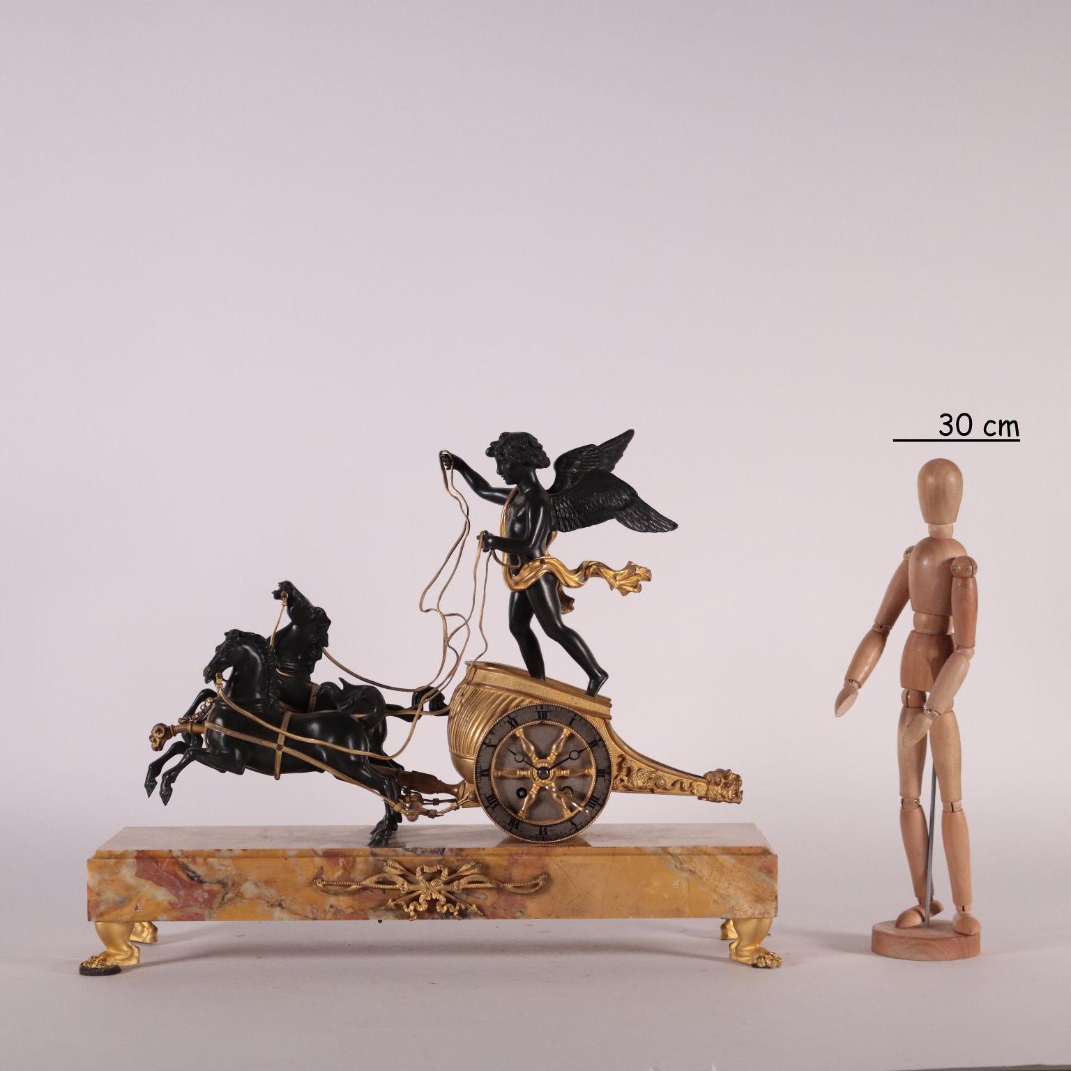 Dark patined gilded bronze table clock with yellow marble of Siena base supported by 4 gilded bronze ferin-shaped feet. The clock is surmounted by winged Cupid on a horse-drawn chariot. Vise with roman numbers for hours and mechanism branded