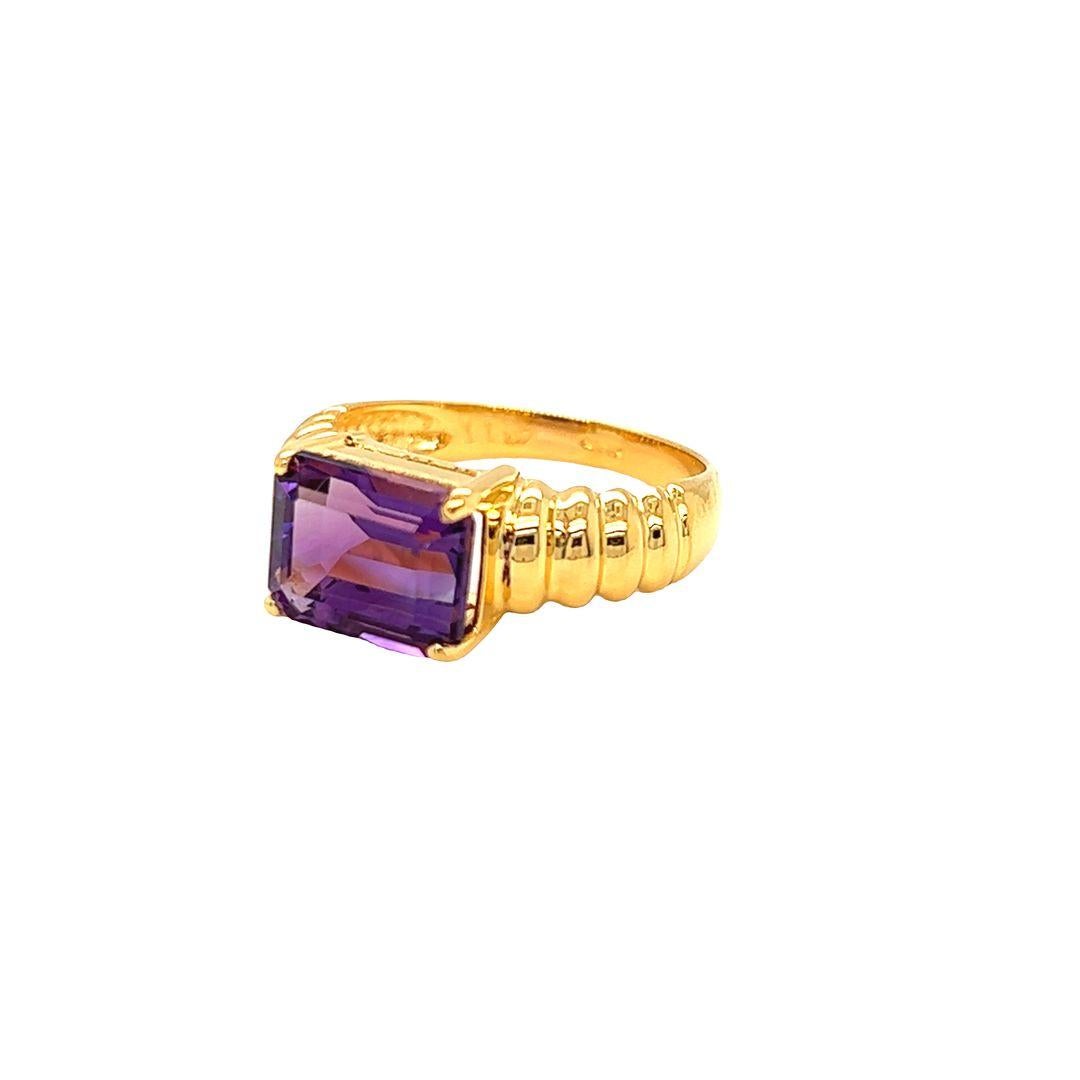 This beautiful Amethyst ring features a classic east-west prong set emerald-cut gemstone, measuring 10 mmx8 mm, and weighing 3 carats. The shoulder of the ring is finished with a textured fluted design and the face of the ring measures 8 mm with a