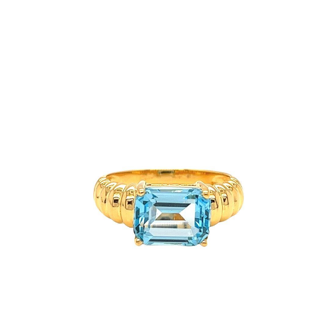 This eye-catching ring features an east-west prong set emerald-cut Blue Topaz that measures 10 mmx8 mm and weighs 3 carats. The ring also has a fluted textured design on the shoulders. The face of the ring measures 8 mm and sits 6.2 mm high off the