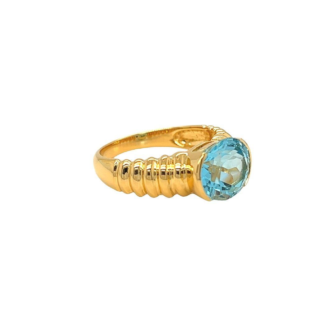 14K Yellow Gold Fashion Ring featuring a stunning east-west half bezel set oval-cut Blue Topaz. The ring showcases a bold and perfectly blue oval Blue Topaz gemstone, measuring approximately 11 mm x 9 mm and weighing 4.5 carats. The semi-bezel