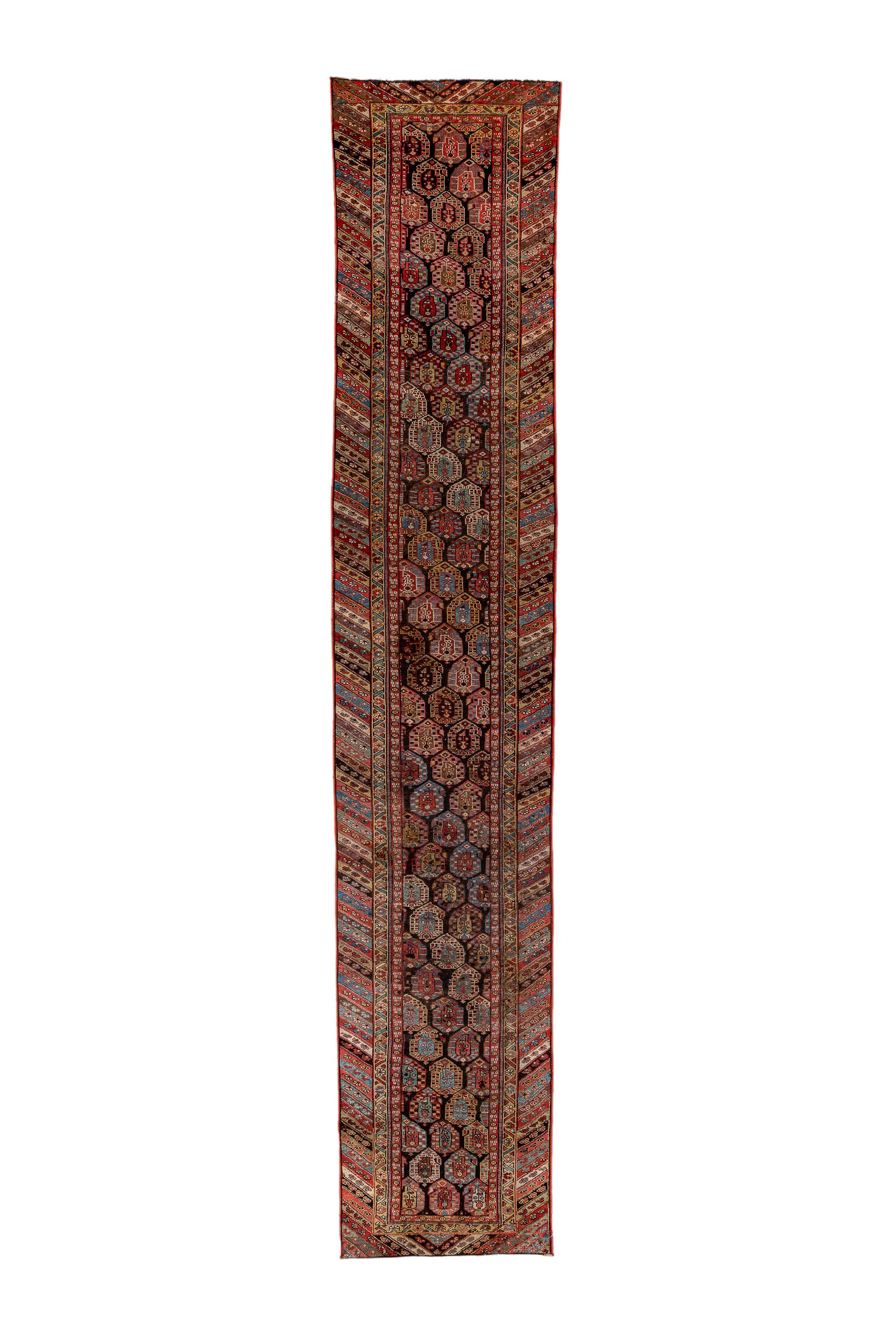 Das fast schwarze Feld zeigt kurze, sich umkehrende Reihen von bunten, geblümten Botehs in einer 2/ 1 plus zwei Hälften halben Tropfen versetzten Reihenfolge. Die Hauptbordüre zeigt diagonale Farbstreifen in Stroh,  hellblau, rot, korallenrot und