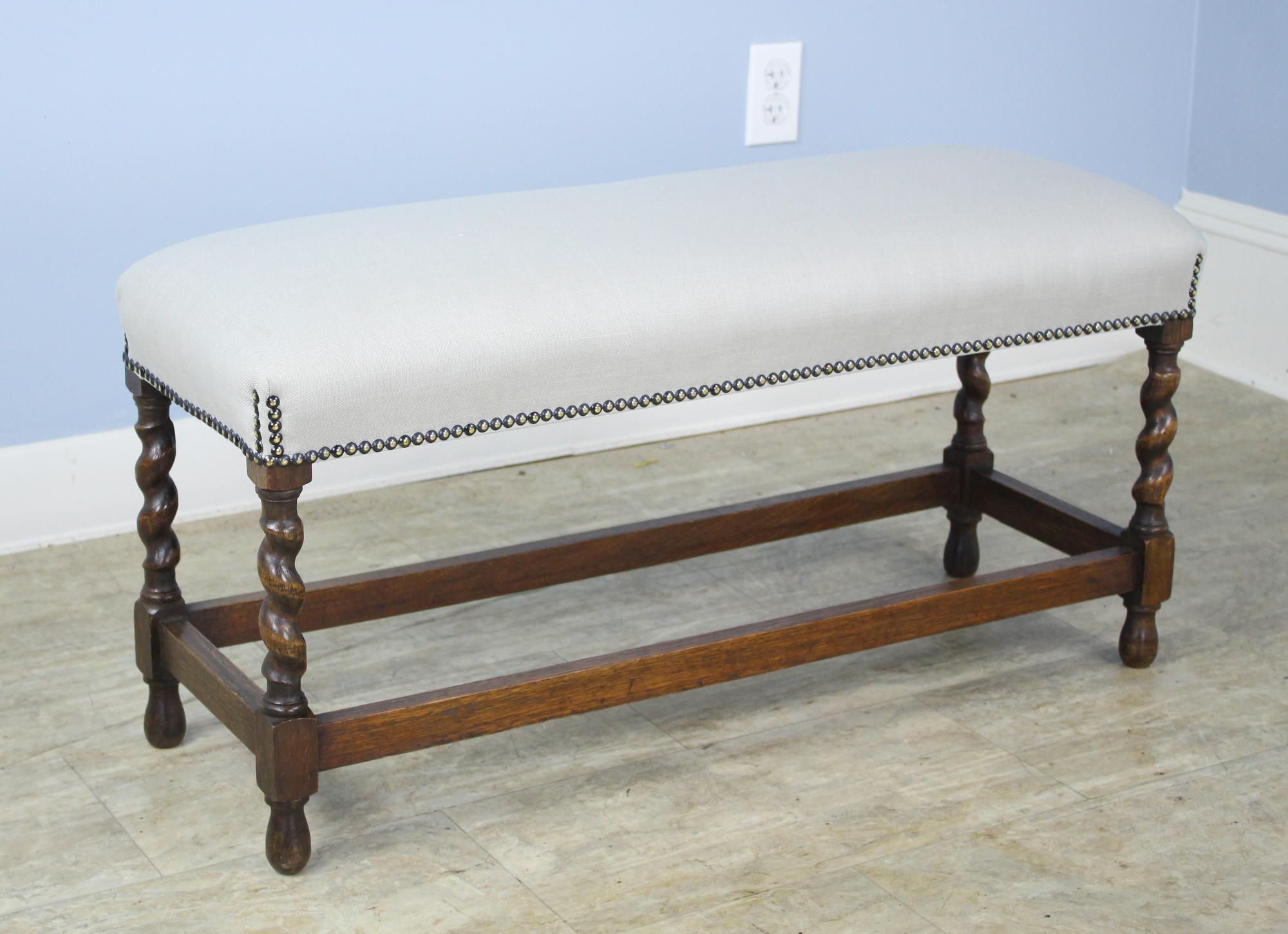 A small stool or bench with visually arresting oak barley twist supports. Good nailhead detail where the seat meets the legs. Newly upholstered in gray linen to be used as is or reupholstered. Charming!