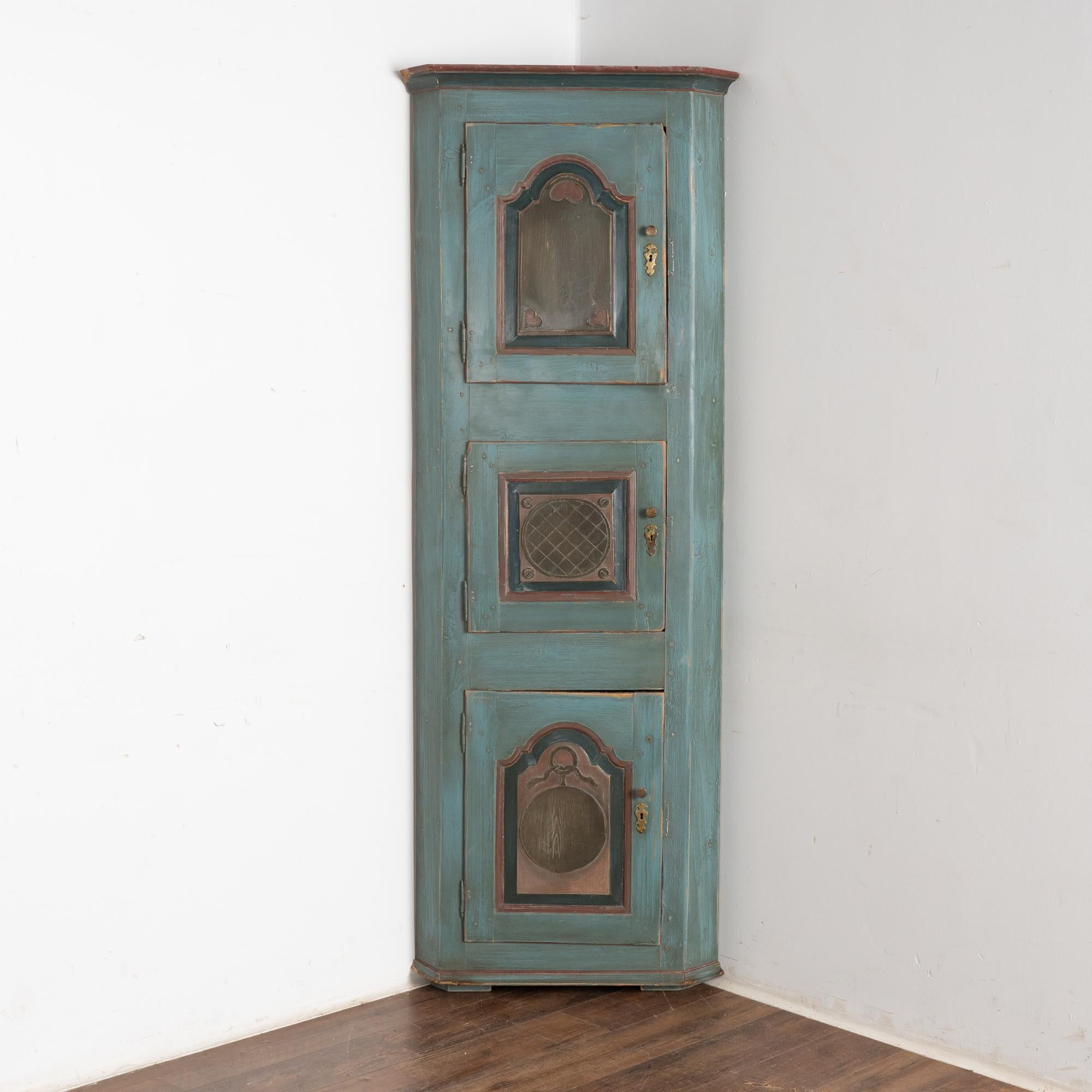 This narrow pine corner cabinet is a special find, due to the original painted finish and unique configuration of the three cabinet doors.
Notice each door panel has a different carved and painted panel, displayed well against the overall blue