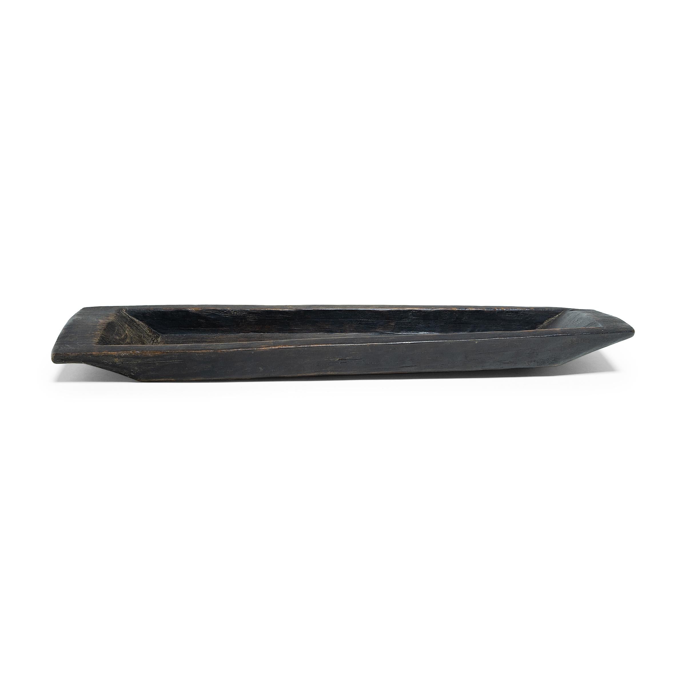 This antique wooden tray perfectly balances minimalist simplicity and rustic charm. Carved from a plank of wood, the tray has a long and narrow form, shaped with a shallow interior, straight sides and tapered ends that flatten into handles. Stained