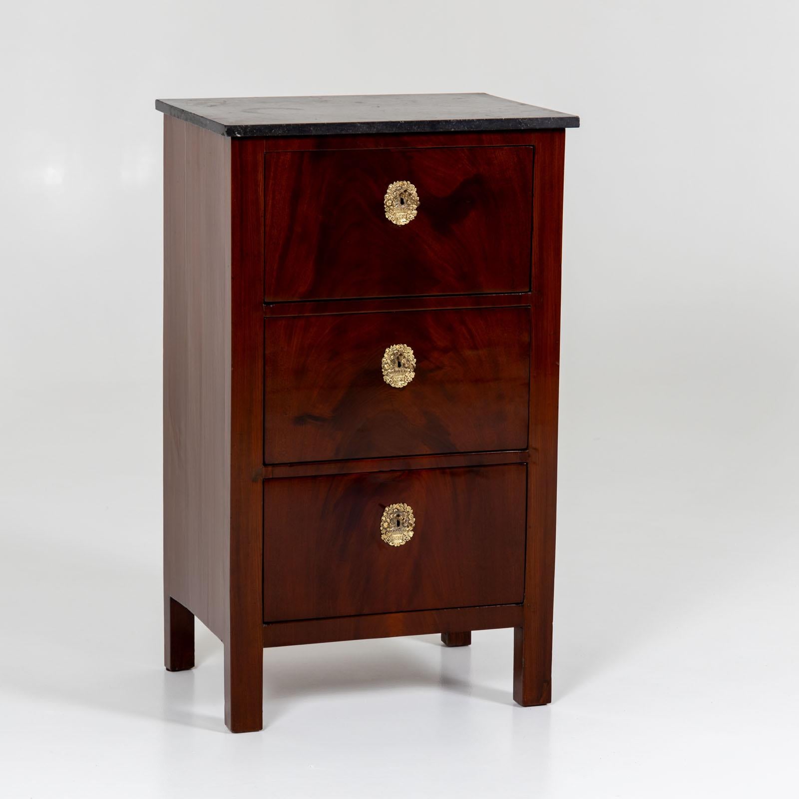 Narrow chest of drawers on straight legs with three drawers and a gray stone top. The key plates are in the form of flower bouquets in gilt bronze. The chest of drawers is mahogany veneered and has been polished by hand. 