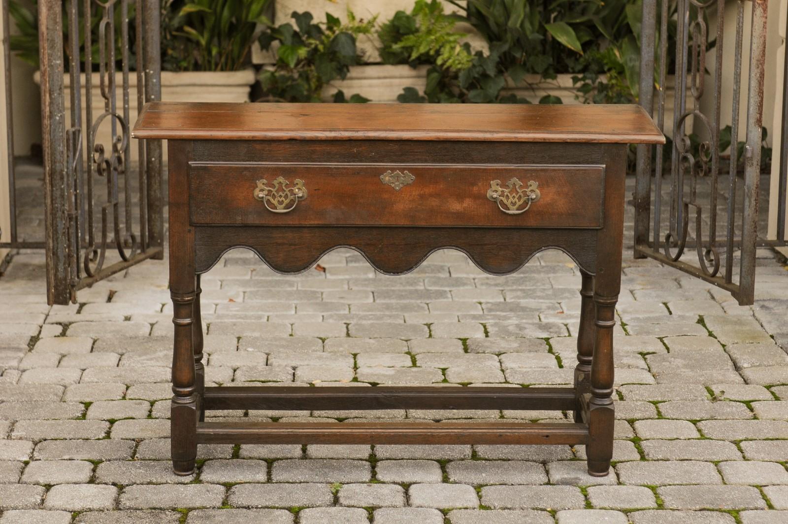 An English narrow oak console table from the late 19th century, with single drawer and scalloped skirt. Born in England during the later years of the 19th century, this elegant console table features a rectangular planked top with rounded edges,