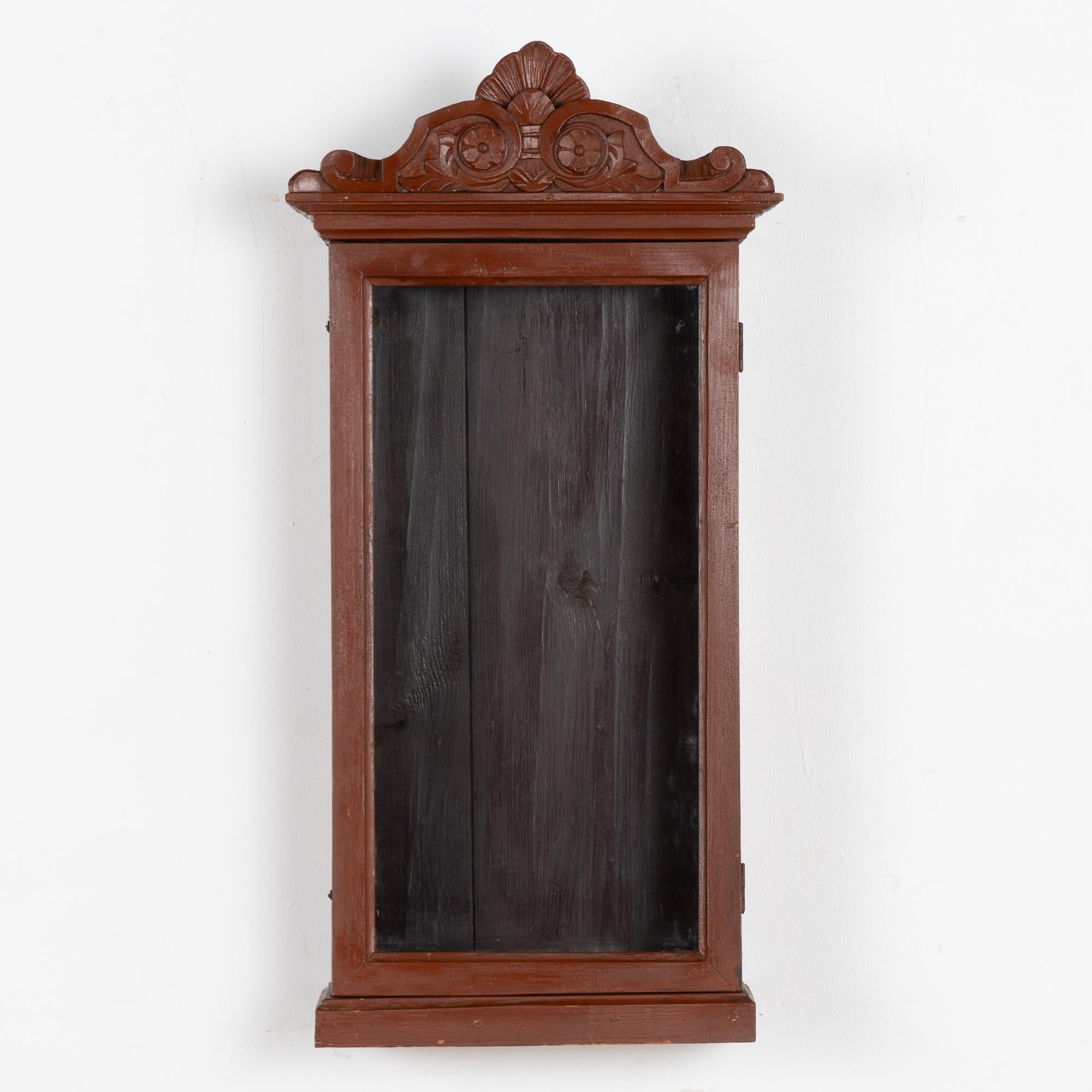 Narrow original brown painted hanging cabinet with decorative carved crown.
Two small hook/latches secure the door which is hinged on the right side. 
All scratches, cracks, dings, scratches and age related separation (see back panel) are reflective