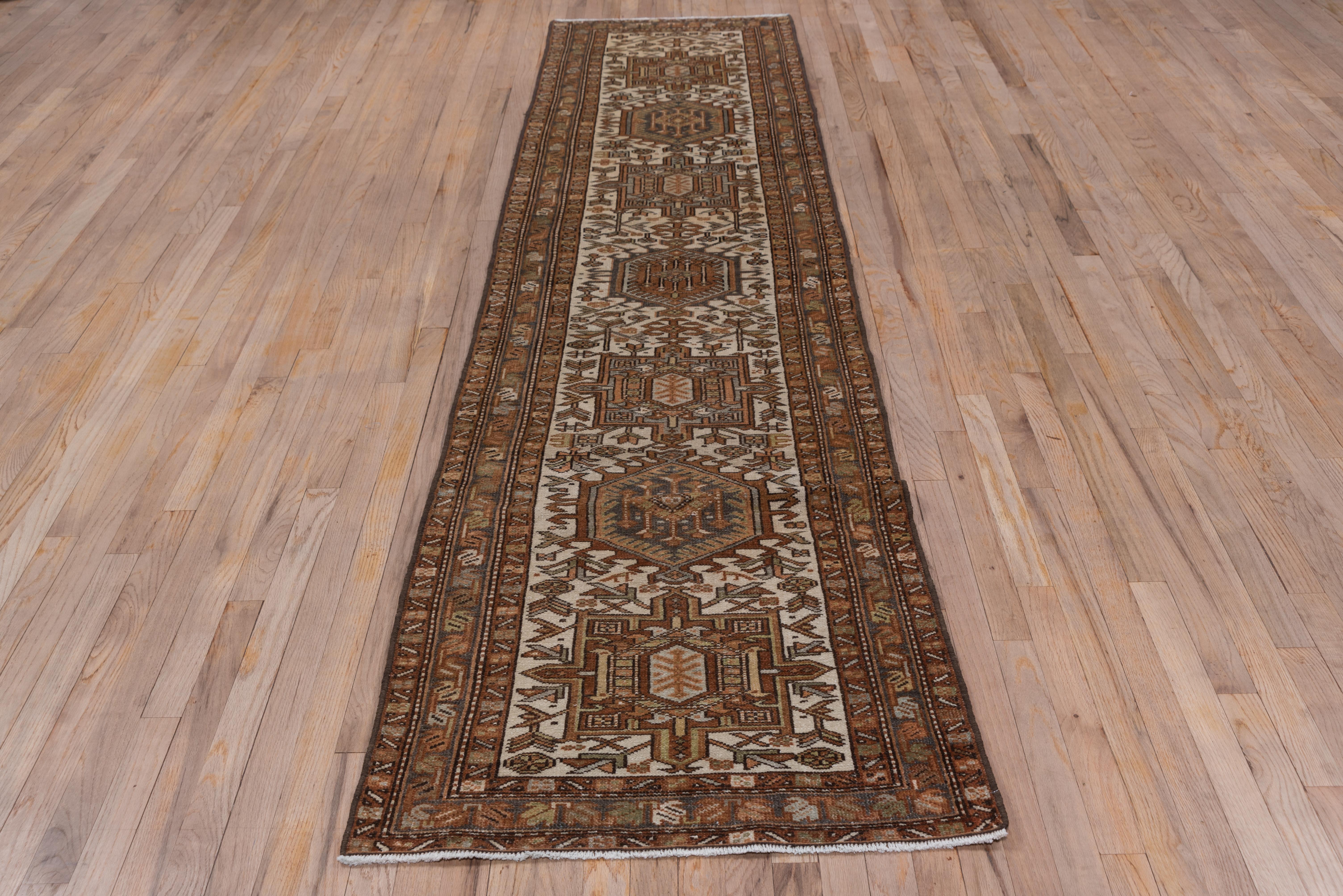 This NW Persian village runner displays the characteristic Karaje octogramme and hooked hexagon medallions, this time detailed in rust on an old ivory ground with stylized flowers filling in. The narrow borders displays squares or a simple
