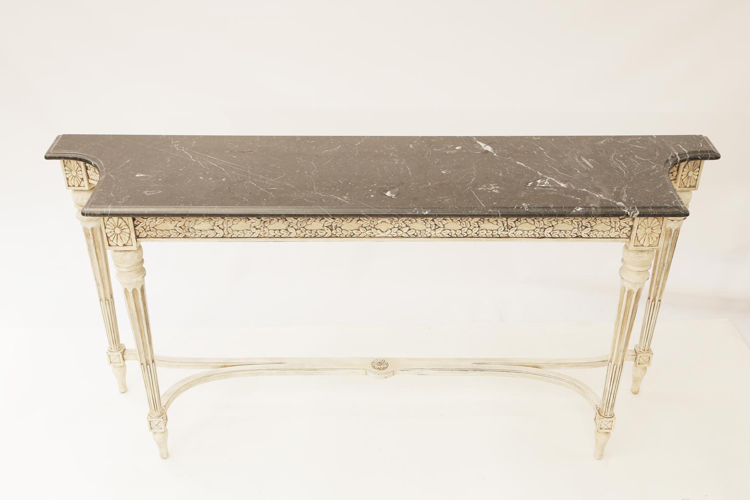 Console table, in the Louis XVI style, having a narrow rectangular top of black Marquina marble with concave, cut-out corners, on conforming table base with a painted finish showing natural wear, its apron carved with laureling, raised on