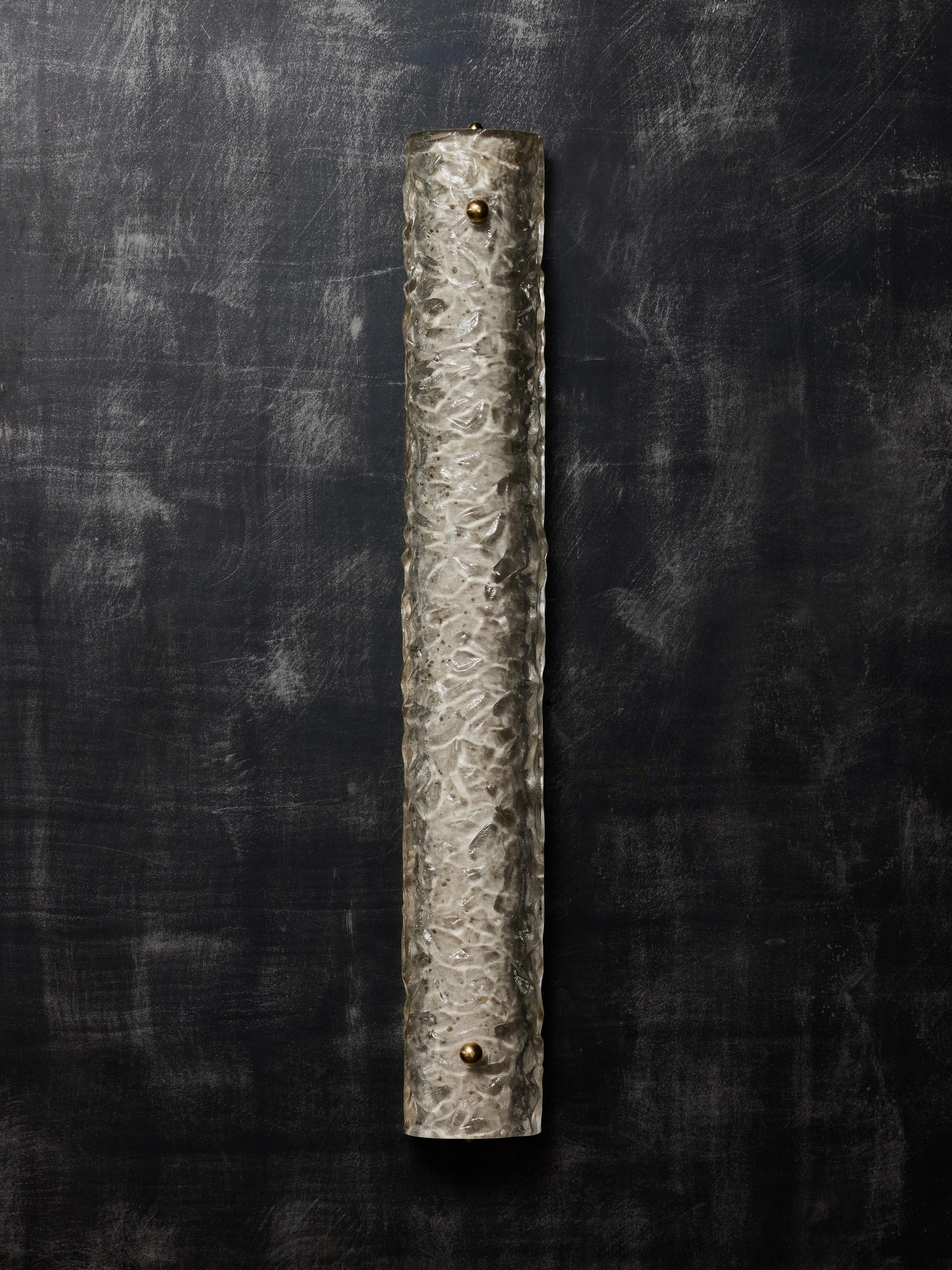 Wandleuchter aus einer Messingplatte, die mit einem gewölbten Murano-Glasstück bedeckt ist, das mit zwei Zierschrauben aus Messing befestigt wird. Der Murano ist strukturiert und hellgrau getönt mit goldenen Reflexen. Drei Lichtquellen pro Leuchte.
