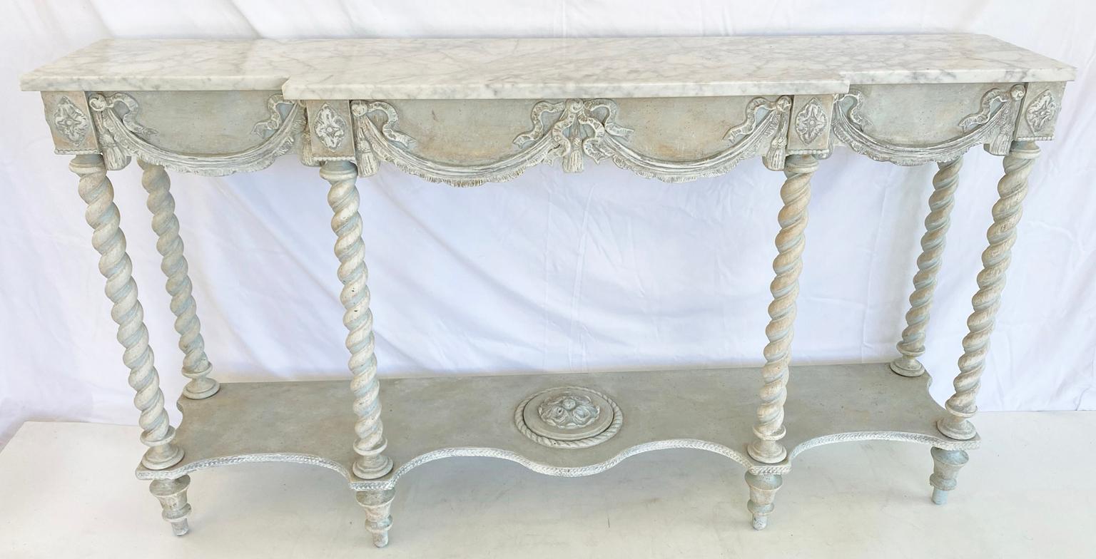 Painted breakfront console table having a top of Carrara marble, on painted table base showing natural wear, its apron carved with swags, ribbons and tassels, raised on six, cartouche-headed, barley-twist legs, joined by a shelf stretcher, centered