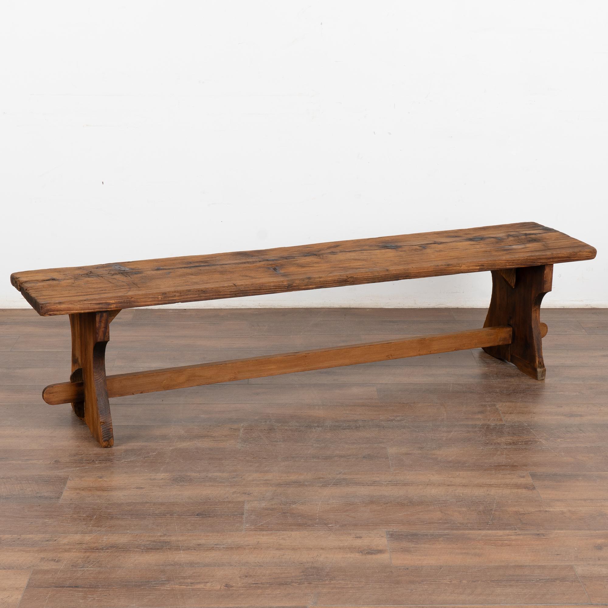 Rustic plank top pine bench with dark aged patina, just under 6' long.
The top reveals generations of use with nicks, gouges, stains, cracks and scuffs which add character to the narrow trestle base bench.
Restored, this bench is strong, stable and