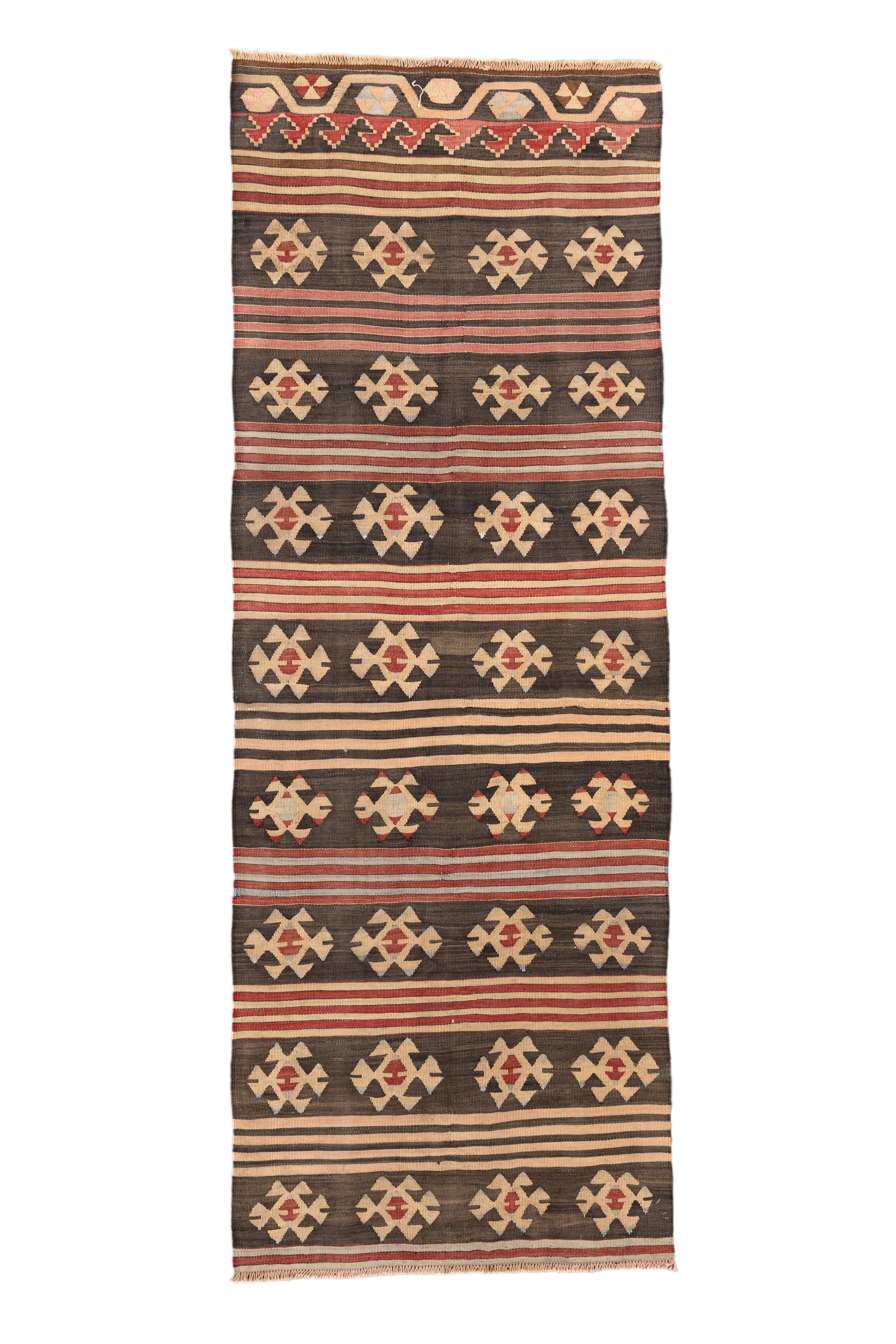  Questo runner Kilim turco vintage combina il minimalismo con l'eredità tribale, con medaglioni ripetuti su un campo marrone intenso e terroso. L'alternanza di file di motivi tribali beige e di strette strisce orizzontali in terra bruciata, ocra e