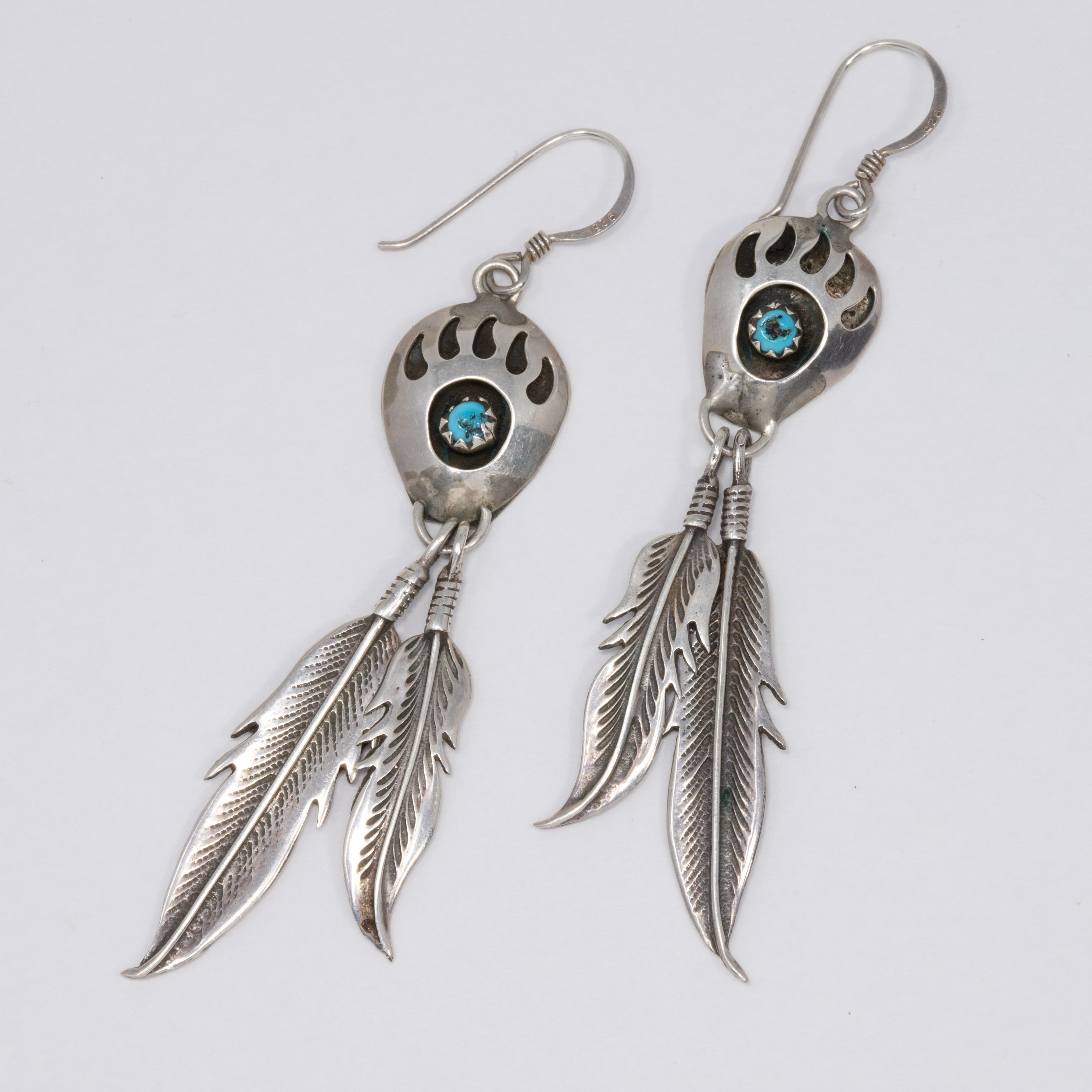 A vintage pair of Native American sterling silver earrings. Each earring features a raised bear claw motif, accented with a turquoise cabochon set in a sawtooth bezel. Two feathers hang off the bottom for a unique touch. Excellent Native American