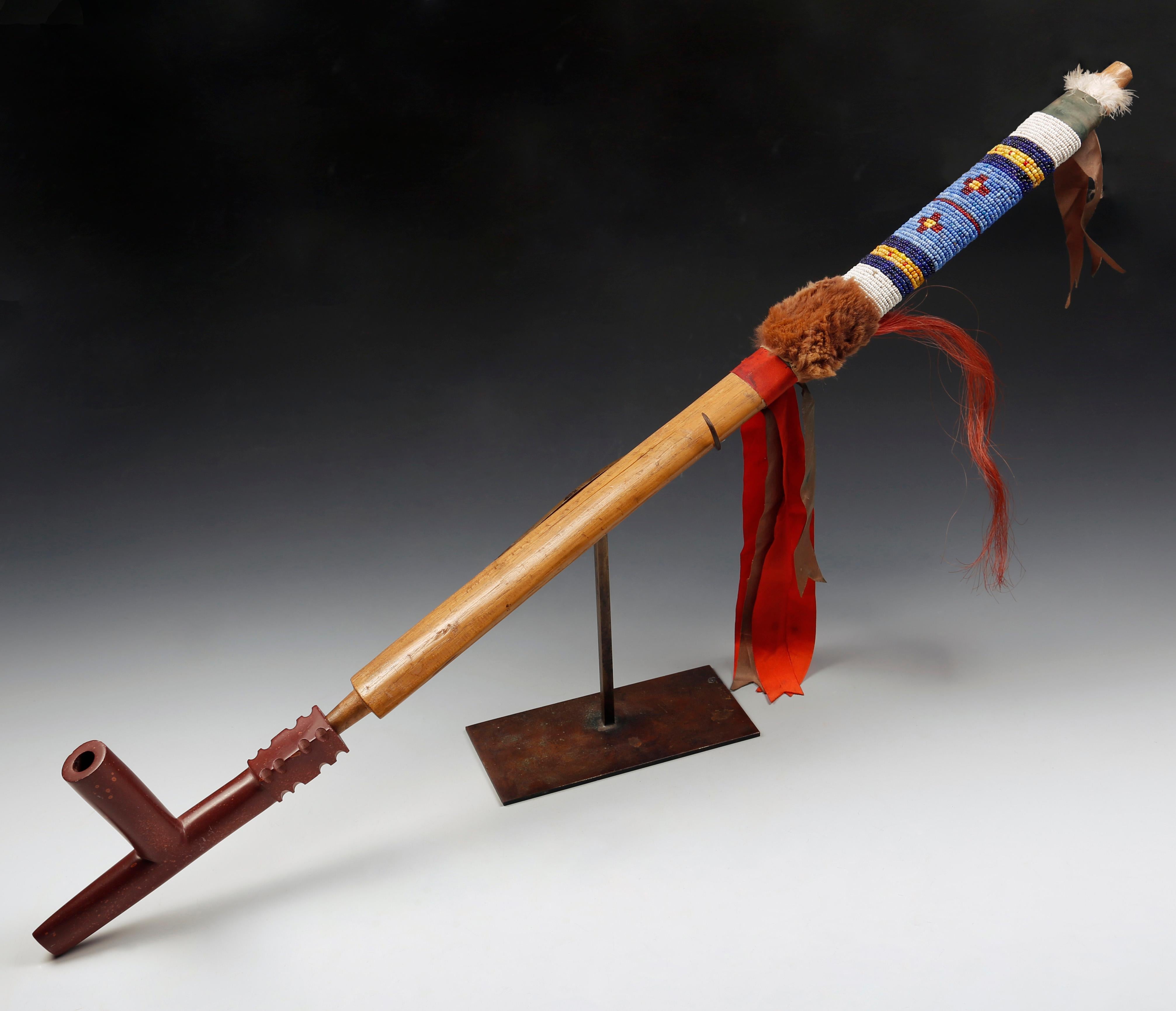 A Native American Indian Sioux beaded wood and caitlinite pipe
A fine Sioux pipe with a T shape catlinite bowl notched block end,
the elliptical wood shaft bound with colored glass beads, fur, feathers, dyed horse hair and ribbons,
81 cm, 32 inches