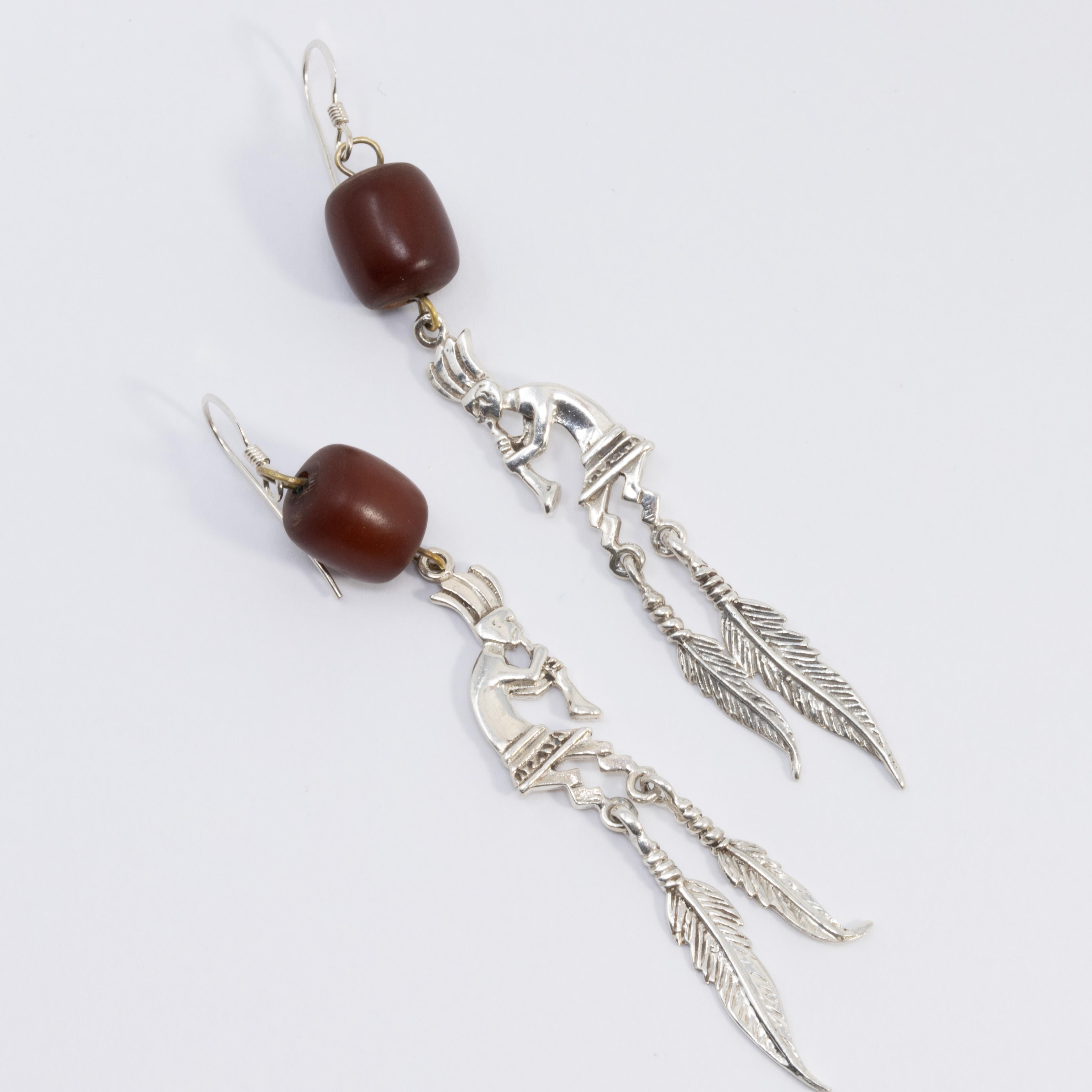 A southwestern style pair of Native American earrings. Features two dangling feathers and a performing horn player, accented with a single ____ bead.

Hook wire hardware