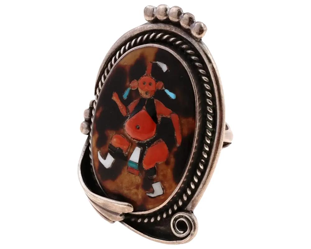 A Southwestern Native American Kachina Silver ring. The ring is made in a medallion shape with a scrollwork design, and adorned with an image of a dancer, encrusted with a Coral stone, and inlaid with multi colored materials. Circa: Mid to the late