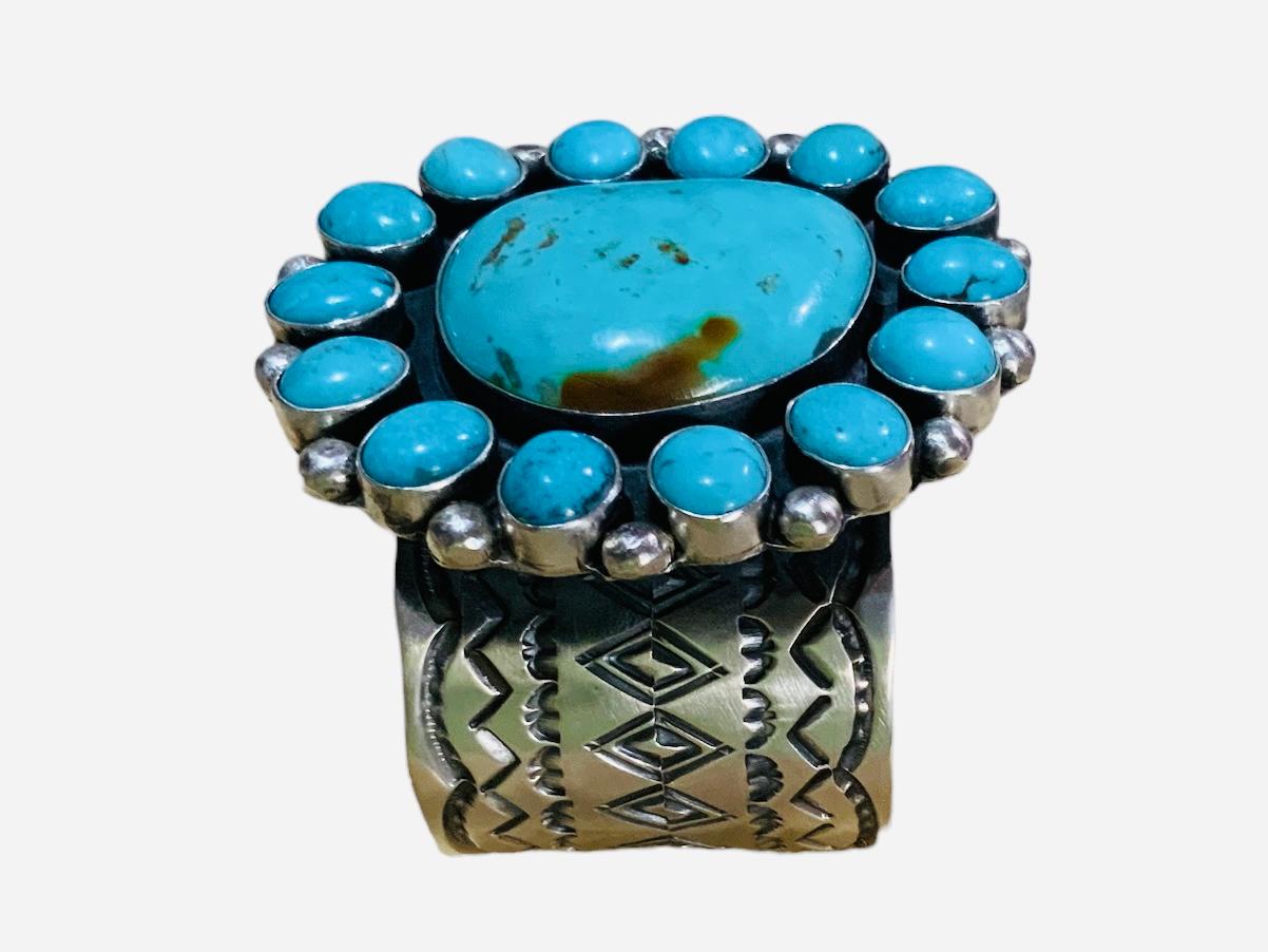 This is a Native American Navajo Sterling Silver and turquoise cuff bracelet. It depicts a wide silver cuff adorned in the front with a “D” shaped like large cabochon turquoise on bezel setting surrounded by fourteen oval shaped cabochon turquoises