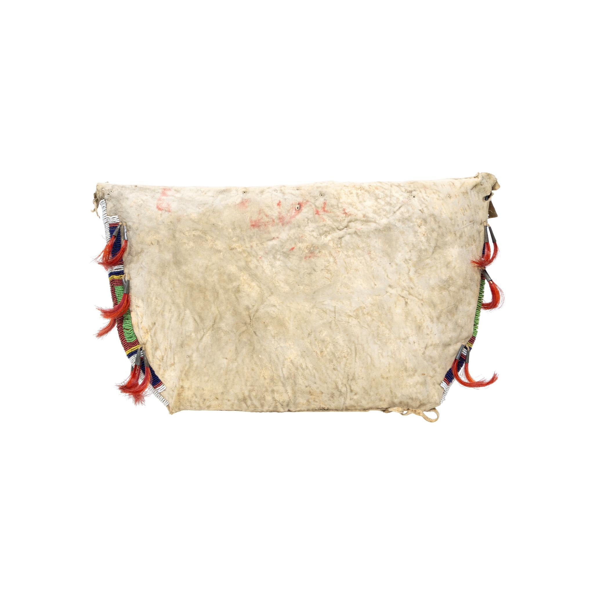 Native American Sioux beaded teepee bag. Having beaded front, sides and top. Beadwork is mostly white with blue, yellow, red, and green forming traditional geometric design. Having tin cones ending in red horsehair against brightly colored beads.