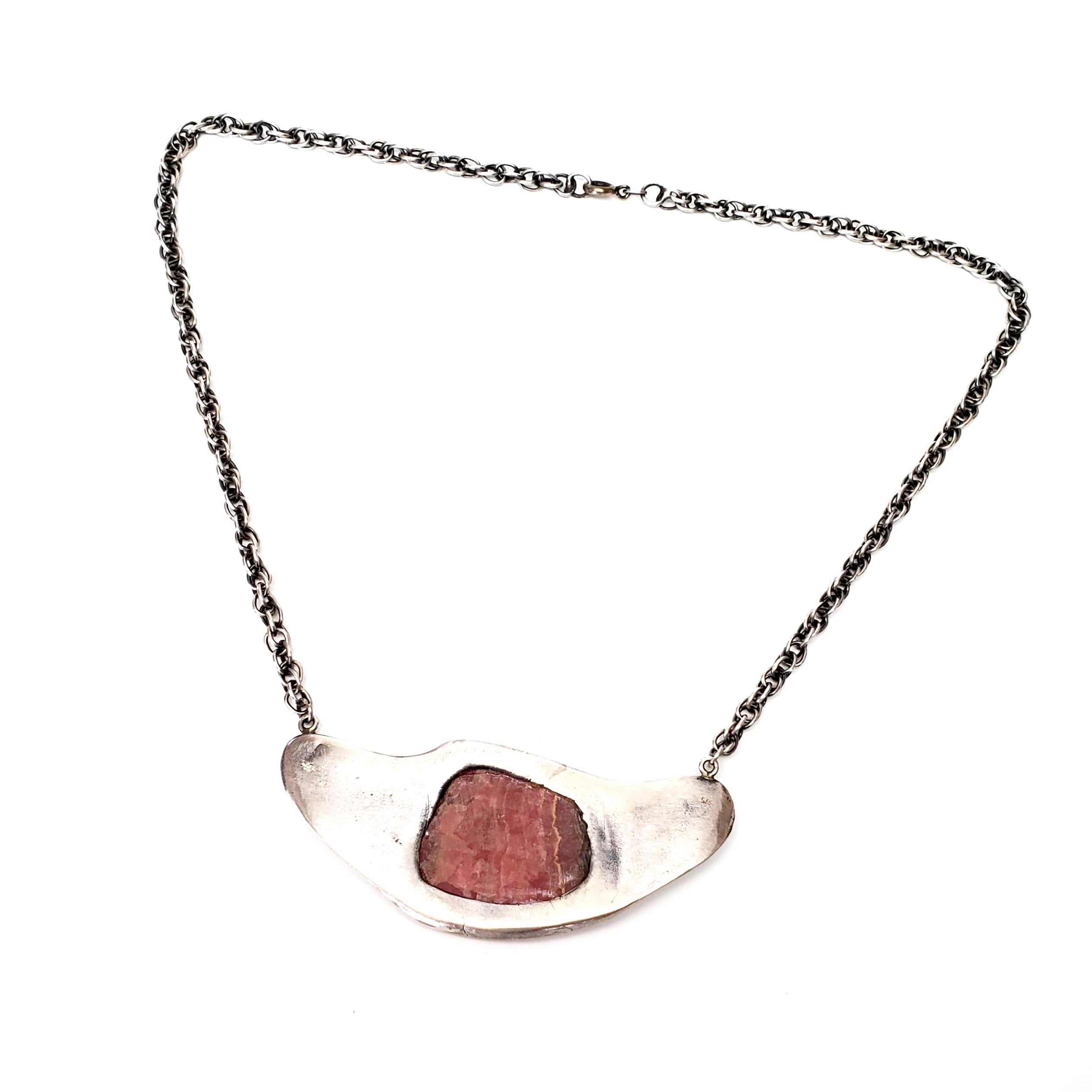 Sterling silver and pink rhodochrosite necklace.

Features a large banded pink rhodochrosite bezel set in a bib featuring a sunny pueblo and boat scene.

Measures approx 19 1/2