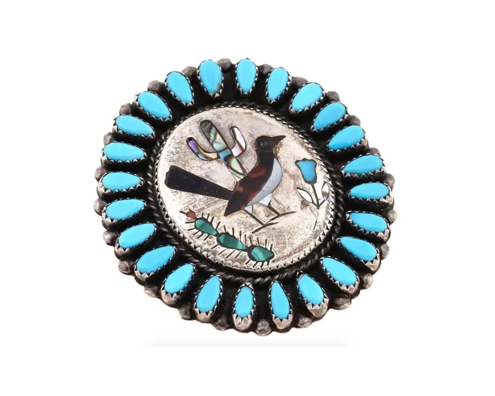 A Native American Zuni medallion shaped Sterling Silver brooch. The brooch is adorned with an image of a bird with a flower and cacti, inlaid with iridescent materials, and encrusted with turquoise stones to the edge. A pin clasp is on the backside.