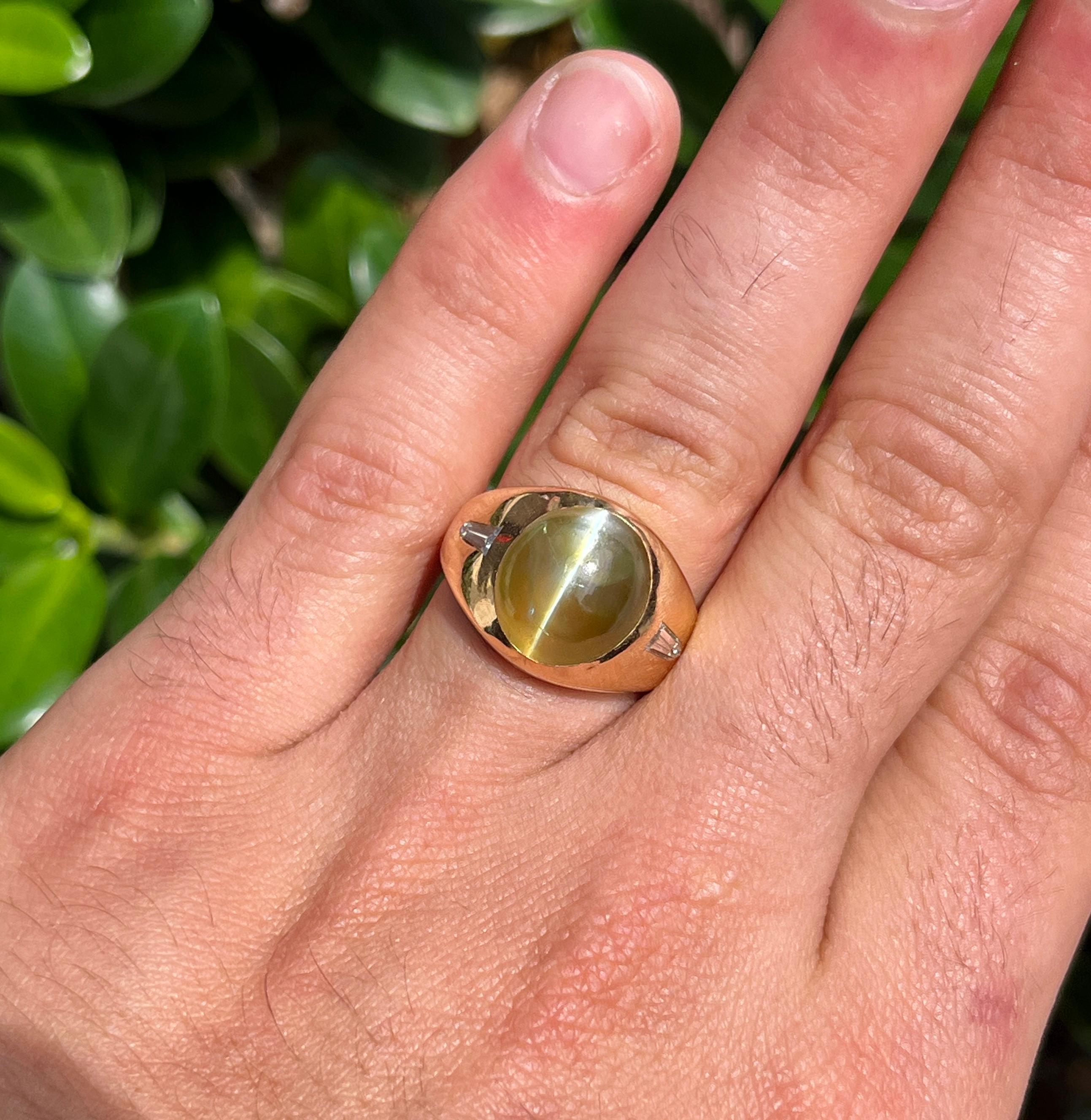 18 karat solid yellow gold men's ring mounts a 12 carat green Chrysoberyl Cat's Eye and tapered baguette diamond side stones. Hand made with a polished finish and secure bezel setting. The center stone bears a remarkable chatoyancy, wit a perfectly
