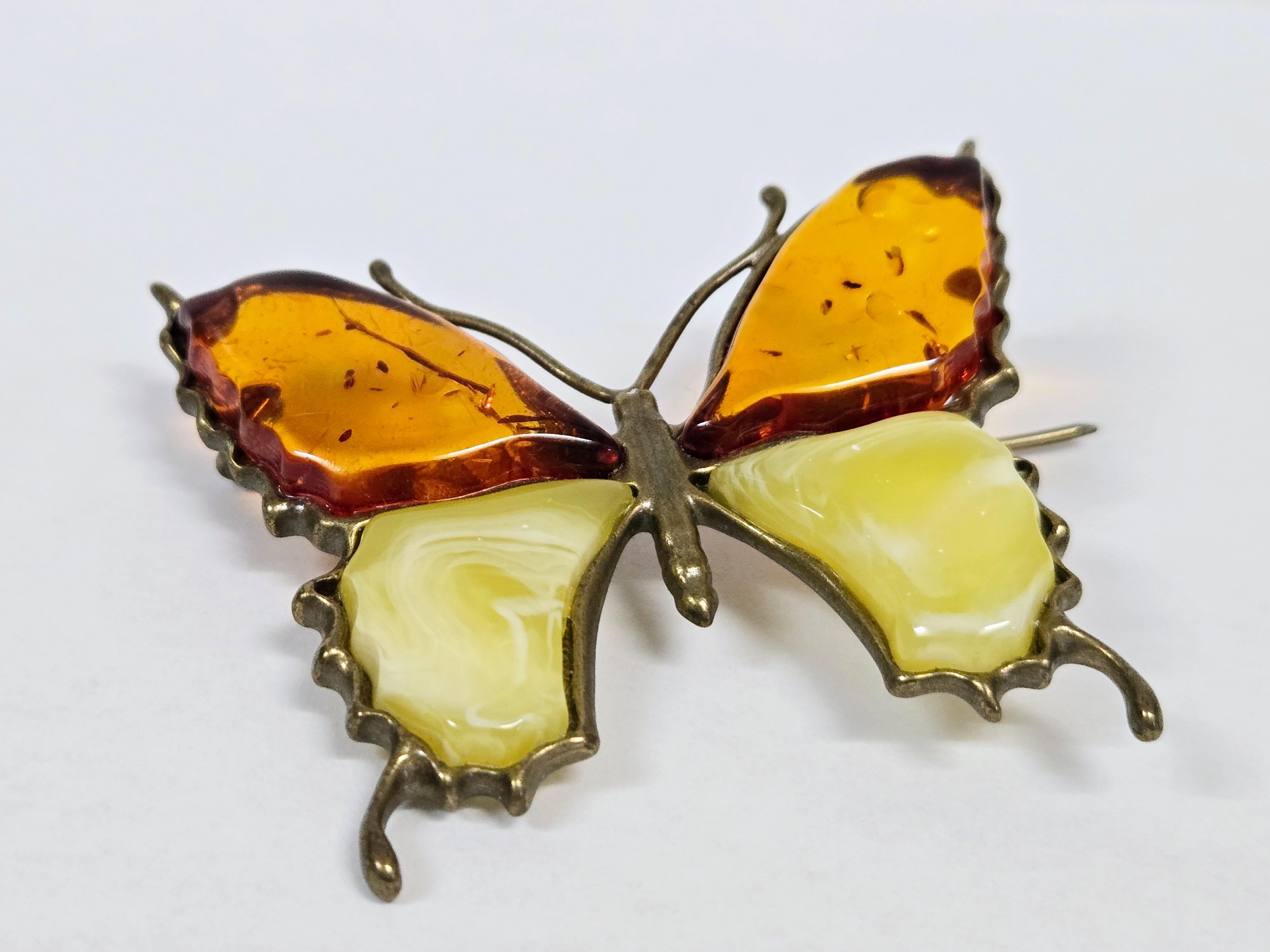 This handcrafted butterfly brooch is made from natural amber, featuring wings in two distinct hues. The upper wings are crafted from classic amber, while the lower wings showcase beeswax amber—a yellow variety with a milky, opalescent appearance and