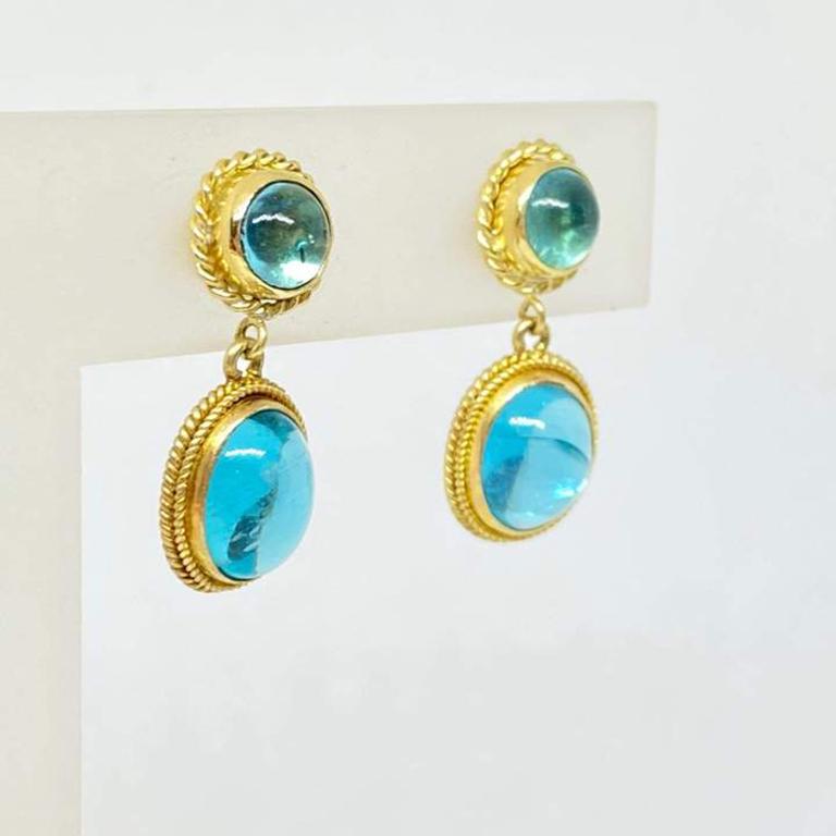 Natural apatite dangle earrings designed in 18K yellow gold. Round bezel set cabochon on top with a woven edge, bezel set oval cabochon dangle with rope edge on the bottom. The earrings have a post and earring back.
