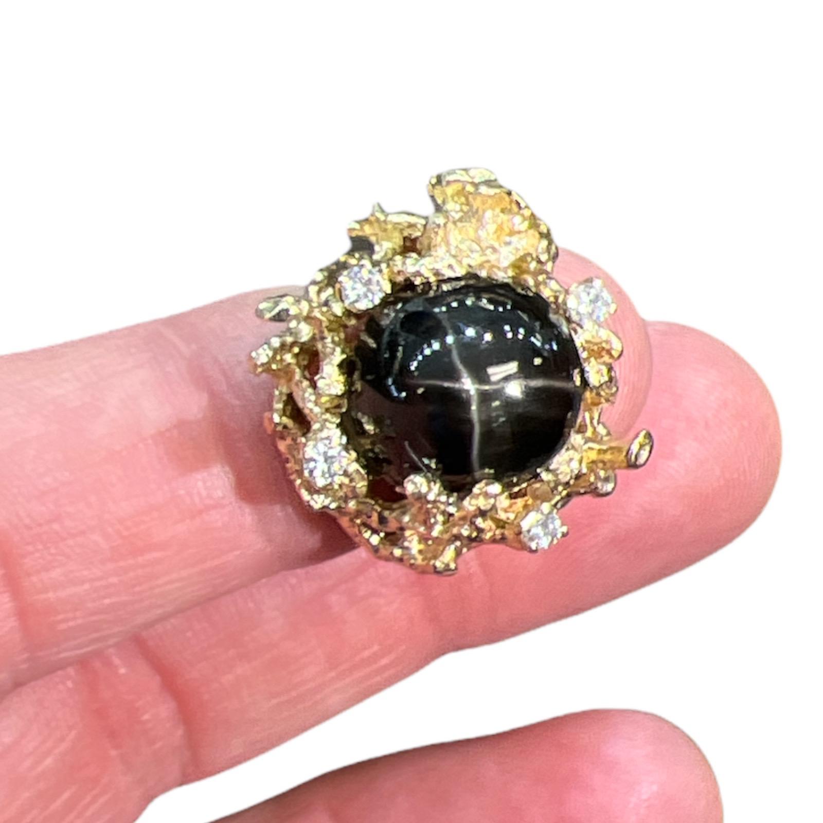 Black star sapphire and diamond ring fashioned in 14 karat yellow gold. The natural black star sapphire is set in an abstract nugget textured ring featuring 4 round brilliant cut diamond accents weighing approximately .12 CTW. The ring measures 20mm