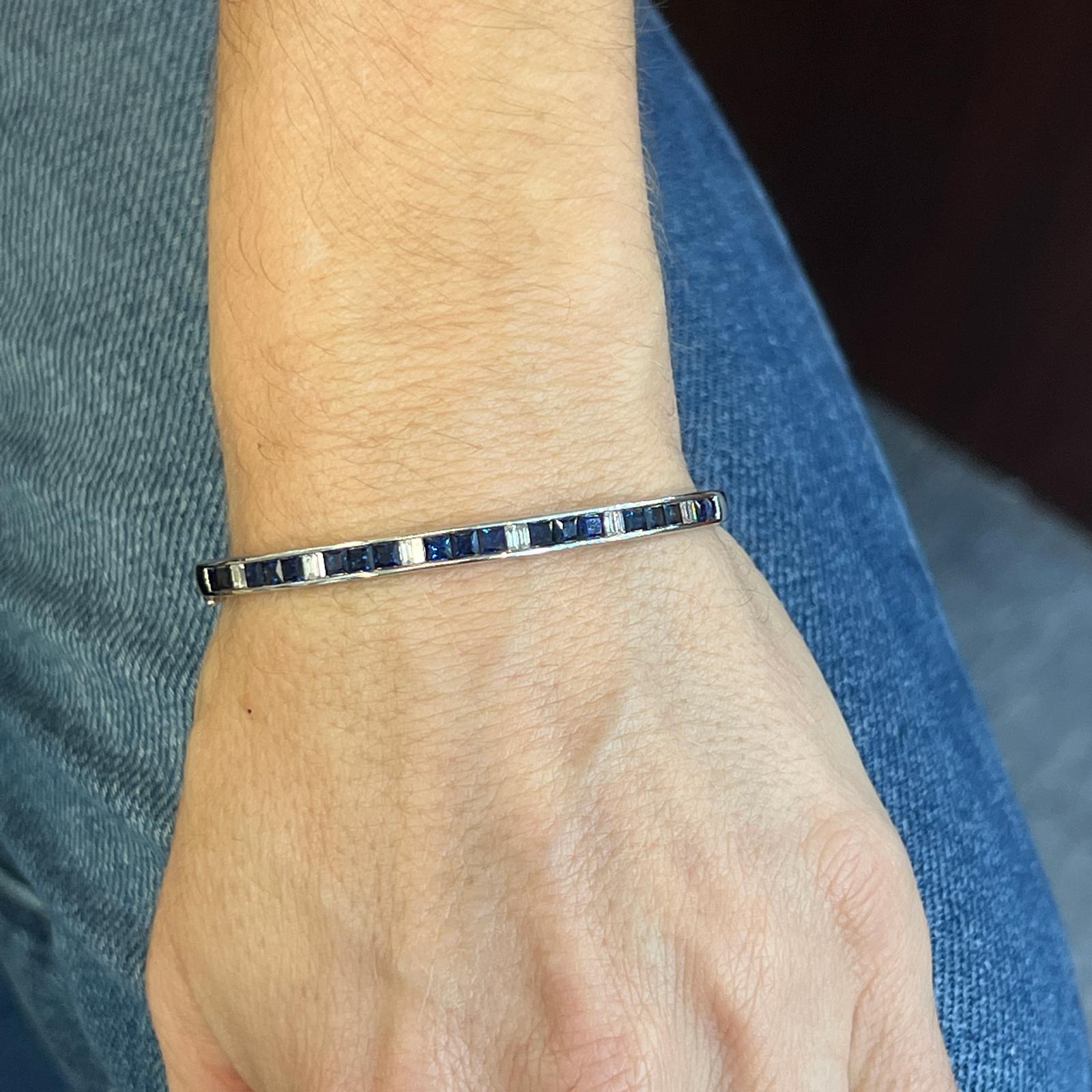 Sapphire and diamond hinged bangle bracelet fashioned in 18 karat white gold. The bangle features natural square cut sapphire gemstones weighing 3.09 carat total weight, and alternating with baguette cut diamonds weighing .36 carat total weight. The