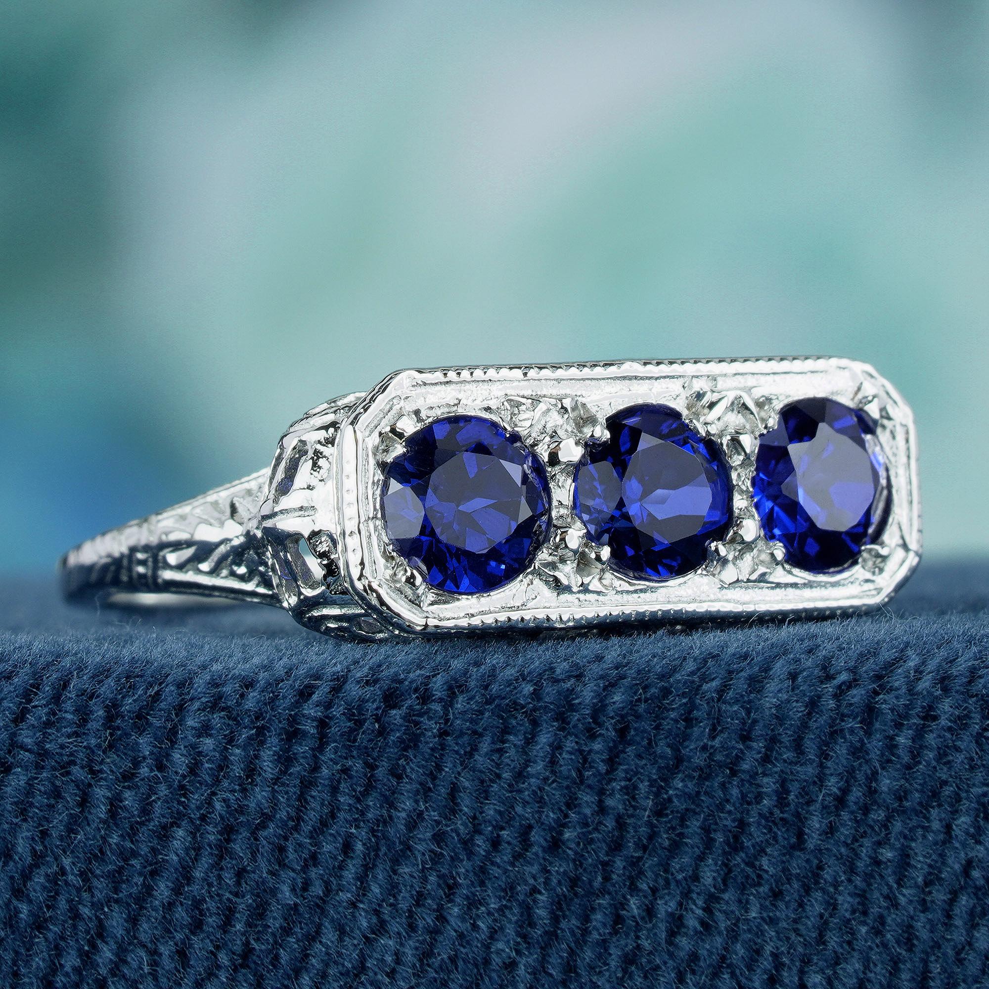 Dazzle with timeless elegance with this exquisite three-stone ring, showcasing mesmerizing round natural blue sapphire gemstones cradled within prongs of a lustrous white gold band. Enhanced with delicate filigree detailing, the ring emanates