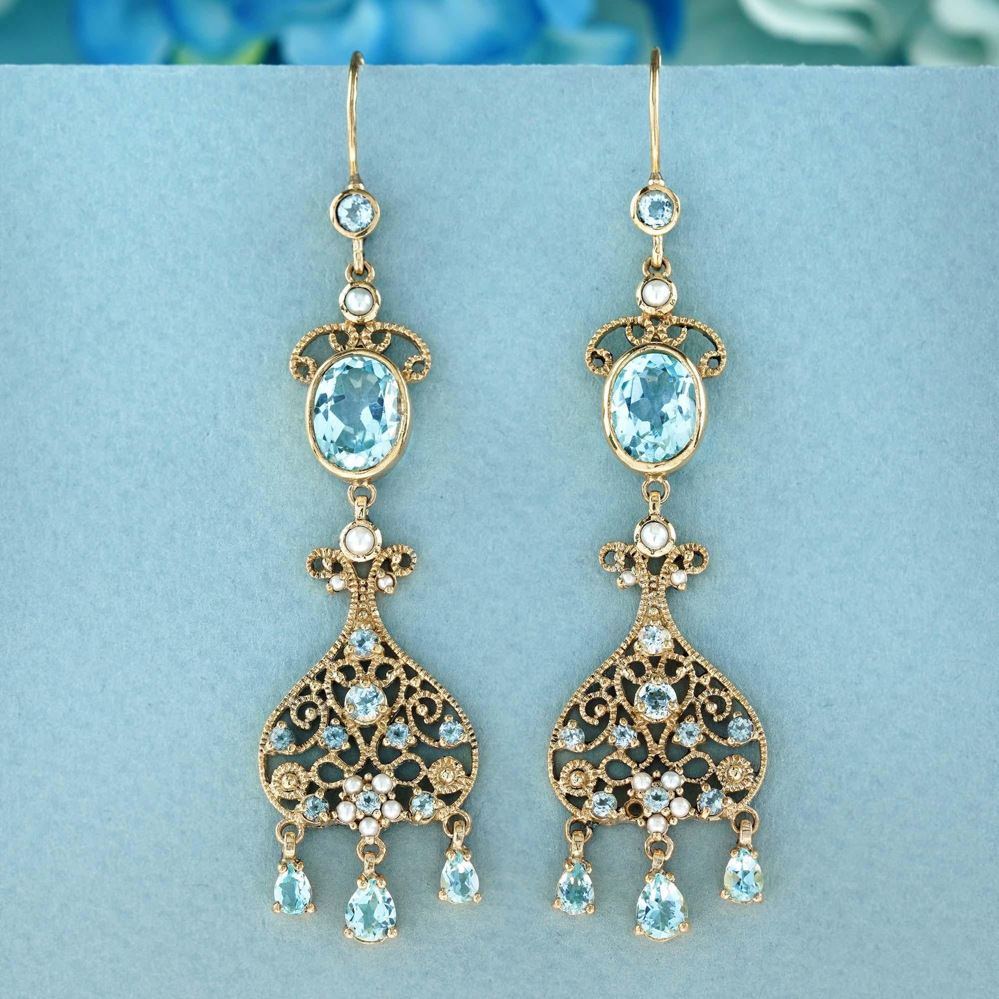 These exquisite vintage-inspired dangle earrings showcase oval-shaped blue topaz gemstones and lustrous pearls, all elegantly set in delicate milgrain yellow gold. The oval-shaped topaz stones exhibit a stunningly clear sky blue hue reminiscent of a