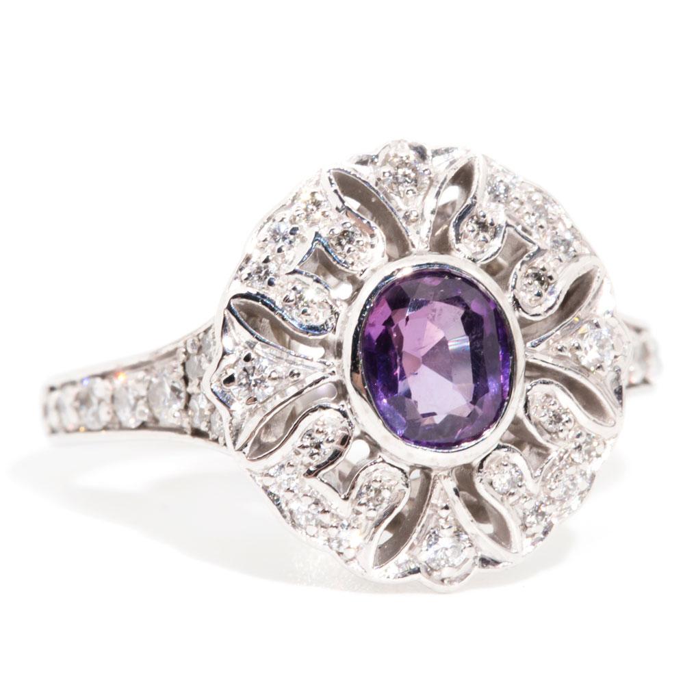 Lovingly crafted in 18 carat white gold, this charming vintage cluster ring features a stunning oval colour shifting sapphire, radiating luminous deep purples and accompanied by glittering round brilliant cut diamonds, adorned around the cluster