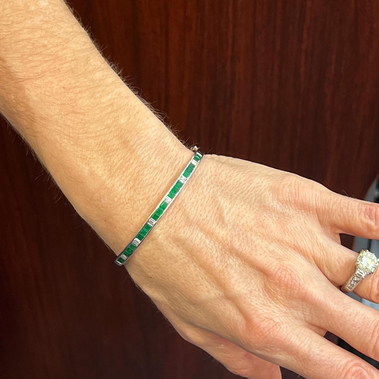 Emerald and diamond hinged bangle bracelet fashioned in 18 karat white gold. The bangle features natural square cut emerald gemstones weighing 2.08 carat total weight, and alternating with baguette cut diamonds weighing .38 carat total weight. The