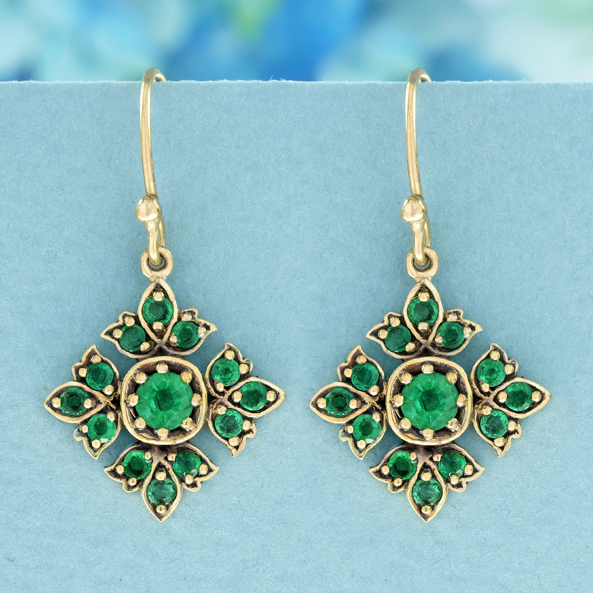These Vintage Style Floral Drop earrings showcase a charming floral design adorned with round, verdant emeralds. Each emerald is delicately set in a prong setting, beautifully accentuating the vibrant green hue against the yellow gold frame. This