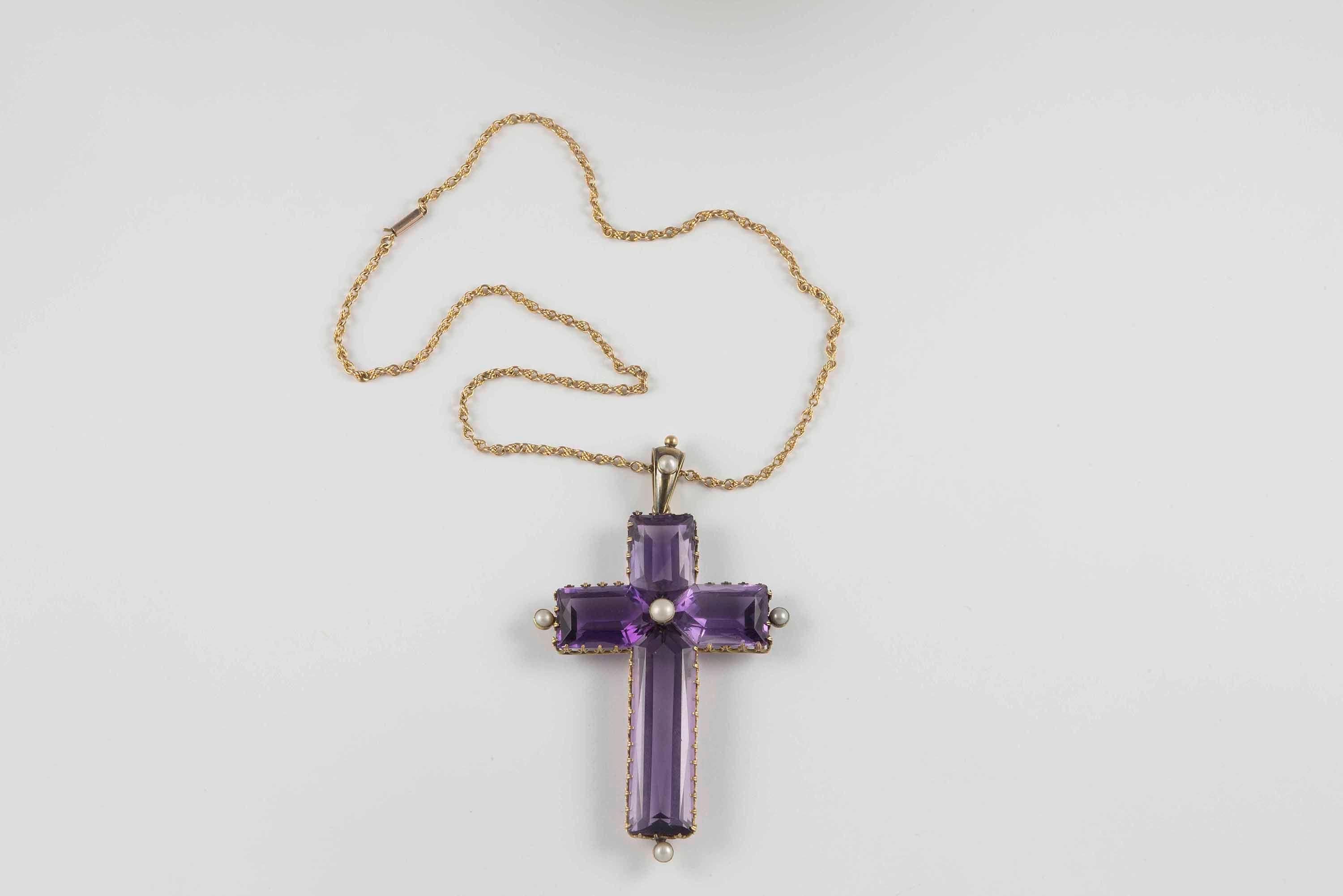 This large antique cross fashioned from 14kt yellow gold is set with a stunning natural fine purple amethyst weighing approximately 100 carats and accented with five seed pearls. The amethyst cross is bordered by delicate handcrafted gold piercing.

