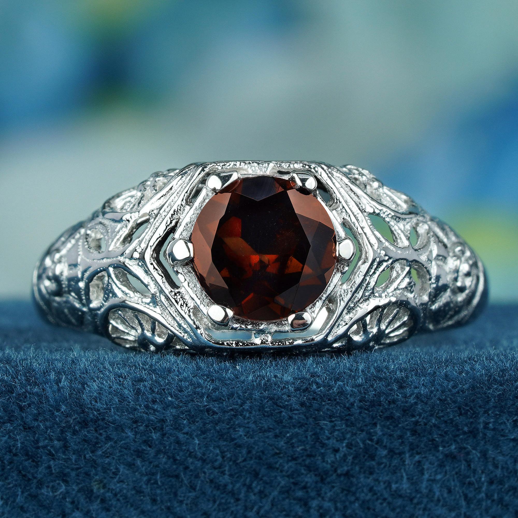 The ring is crafted from solid white gold, giving it a cool, timeless feel. The wide, flat shank tapers gently towards the red garnet in a prong setting, adding a touch of elegance and proportion. The intricate floral design that graces the entire