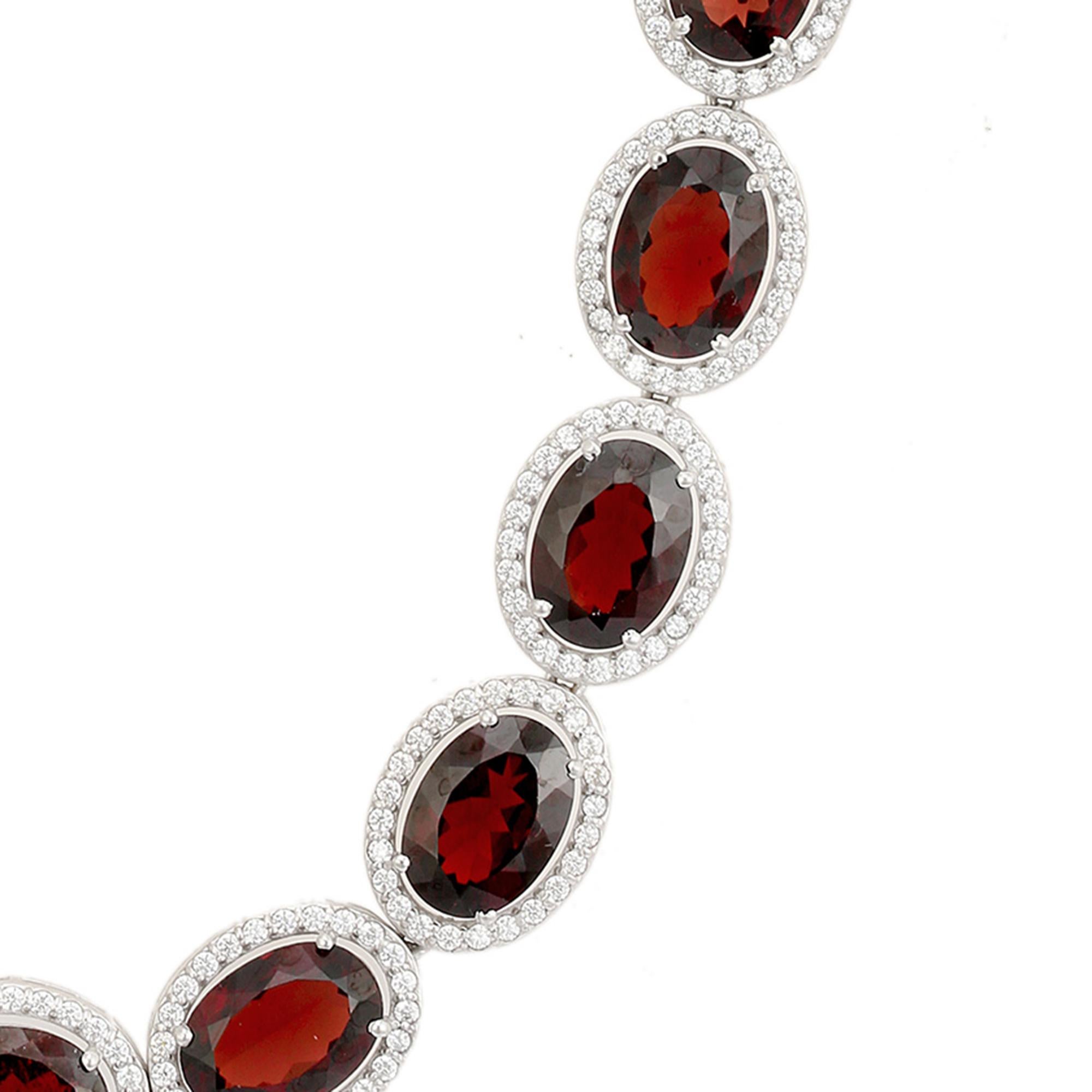 Ce collier ras du cou en grenat naturel et zircon, serti à la main, présente une série captivante de grenats ovales d'un rouge profond, chacun entouré d'un halo lumineux de topaze blanche. Serti en argent sterling 925, le design allie sophistication