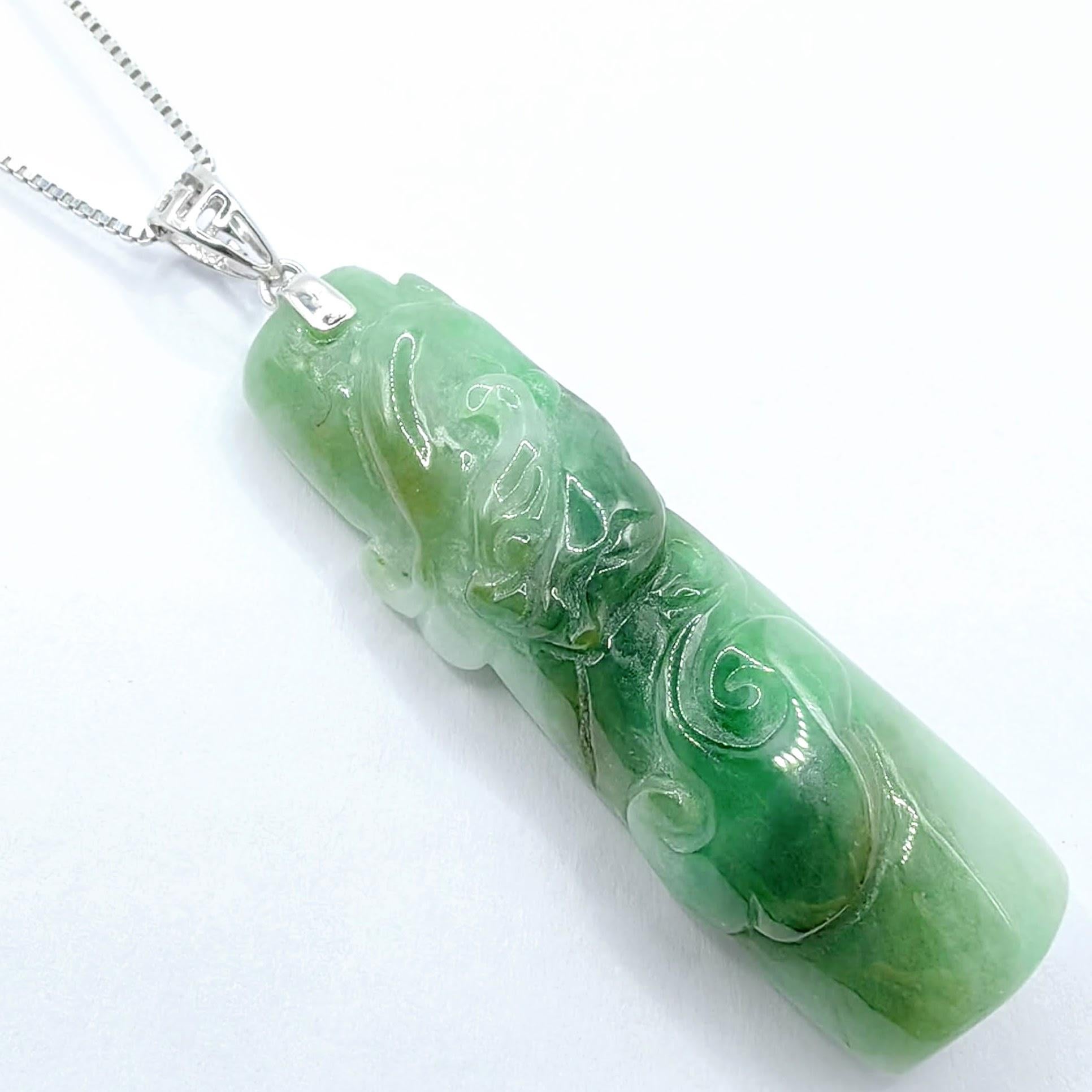 This striking pendant features a stylistic carving of a traditional Chinese dragon, artfully rendered in a natural green jadeite. The slightly translucent jade exhibits soft medium green tones with gentle cloud-like mottling and natural color