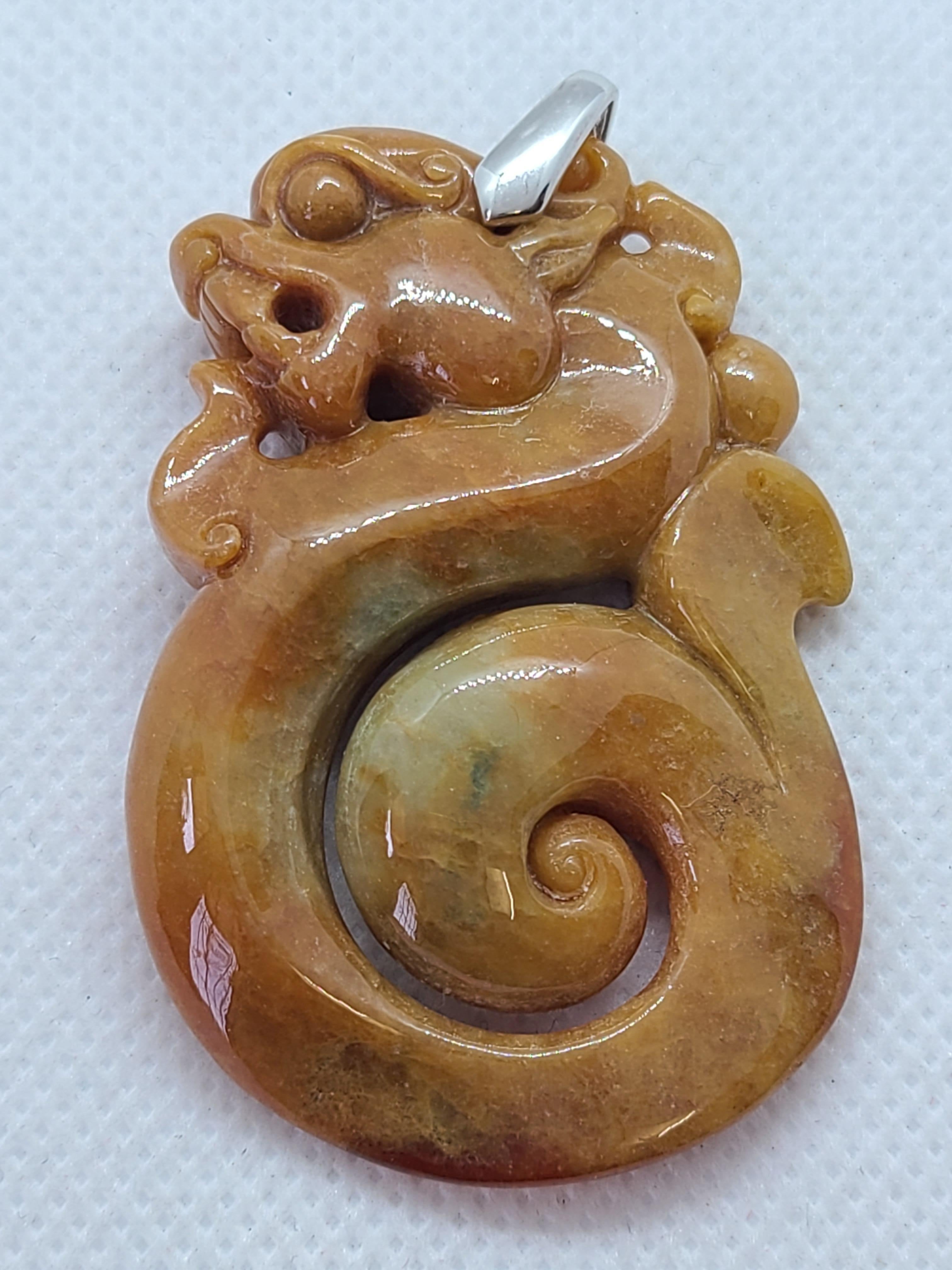Questo ciondolo mostra una miscela naturale di marrone miele chiaro e ricco con accenni di giada verde grigiastra, sapientemente scolpita a forma di antico drago. Il ciondolo misura circa 37,05 x 55,53 x 4 mm e viene fornito con un rapporto che ne