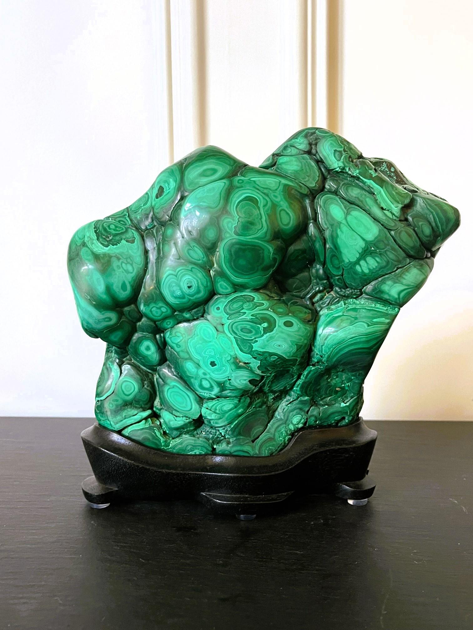 A natural malachite rock specimen with striking green and black colors fitted on a wood stand for viewing and meditating. The front and sides of the gemstone feature tight and small polished botryoidal form, revealing a beautiful, mottled pattern