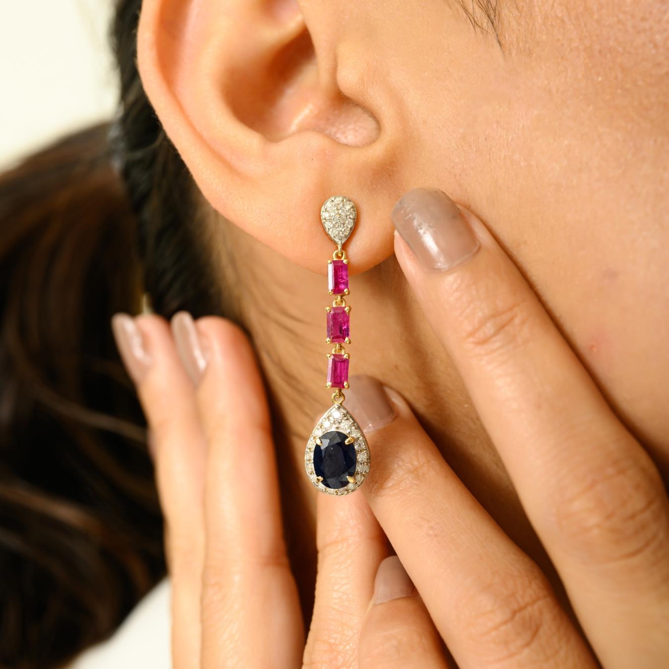 Combinaison saisissante de couleurs vives et d'élégance intemporelle, ces boucles d'oreilles pendantes en or jaune 14 carats sont ornées de rubis éclatants (4,31 cts) disposés en une gracieuse ligne verticale. À la base, des saphirs d'un bleu