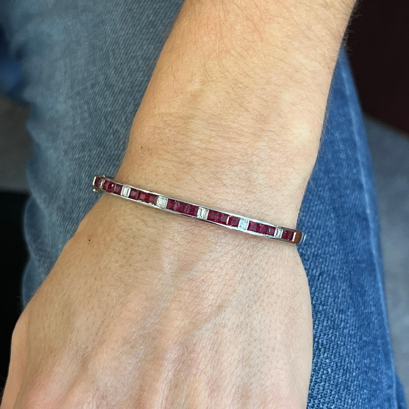 Ruby and diamond hinged bangle bracelet fashioned in 18 karat white gold. The bangle features natural square cut ruby gemstones weighing 2.98 carat total weight, and alternating with baguette cut diamonds weighing .45 carat total weight. The