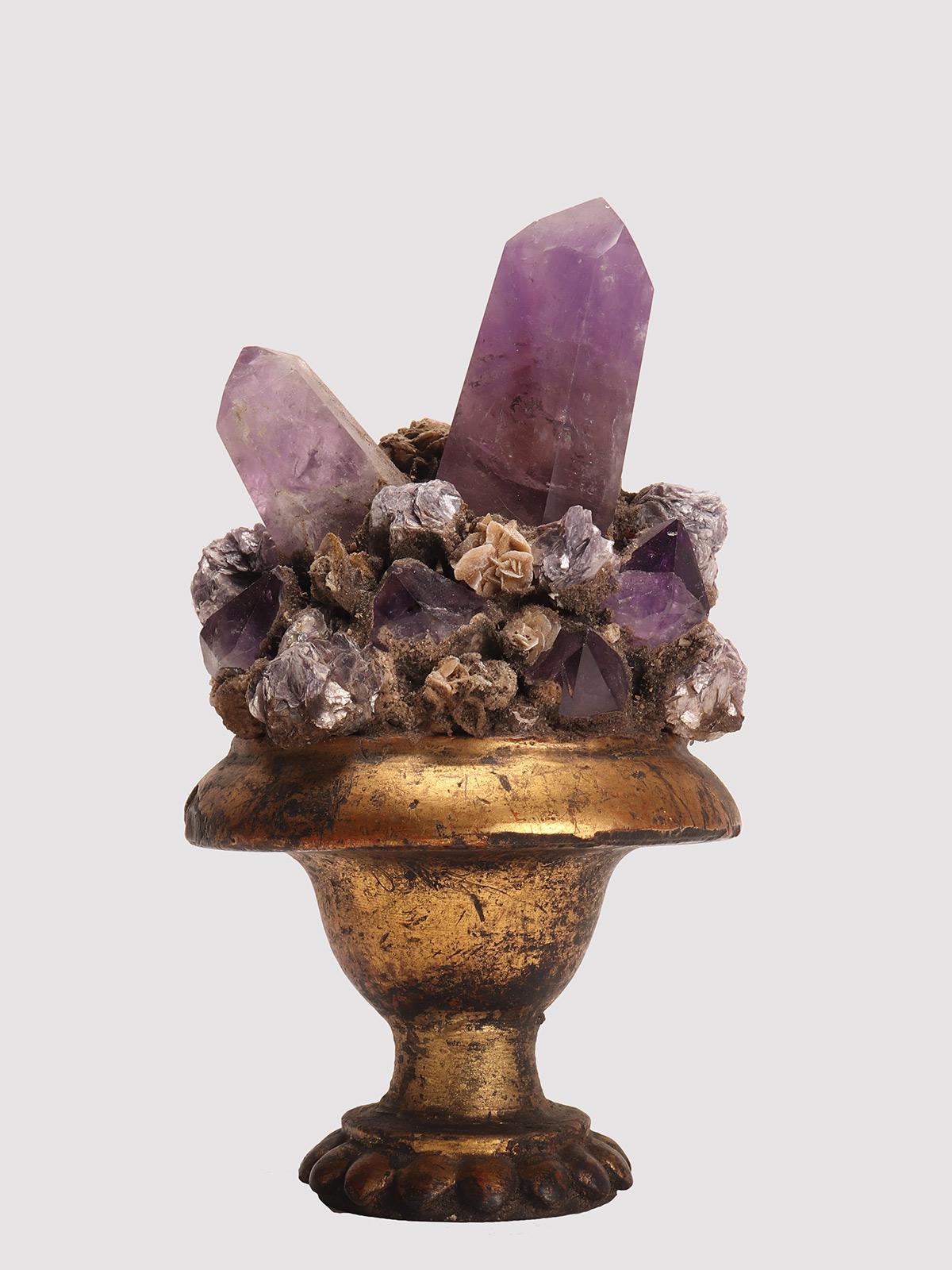 Un ejemplar de mineral de Naturalia. Drusa con cristales de amatista, mica y rosa del desierto, montada sobre una base de madera dorada en forma de jarrón. Italia, hacia 1880.