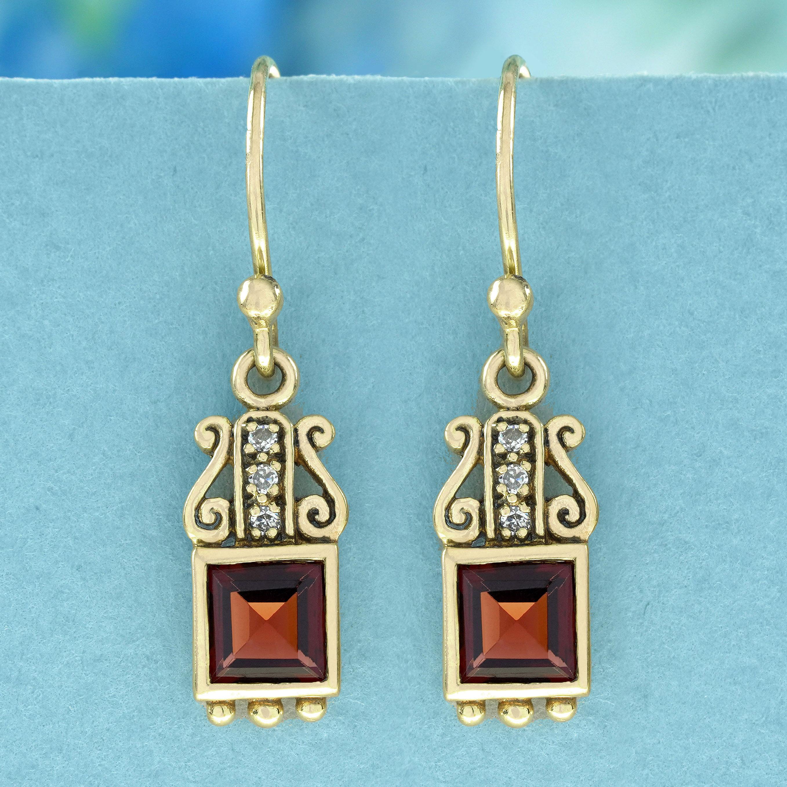 Crafted in vintage-style drop design, these yellow gold earrings feature a deep red square-cut garnet at the bottom. Adorning the top are three cascading round diamonds, offering a touch of sparkle, complemented by delicate scrollwork, culminating