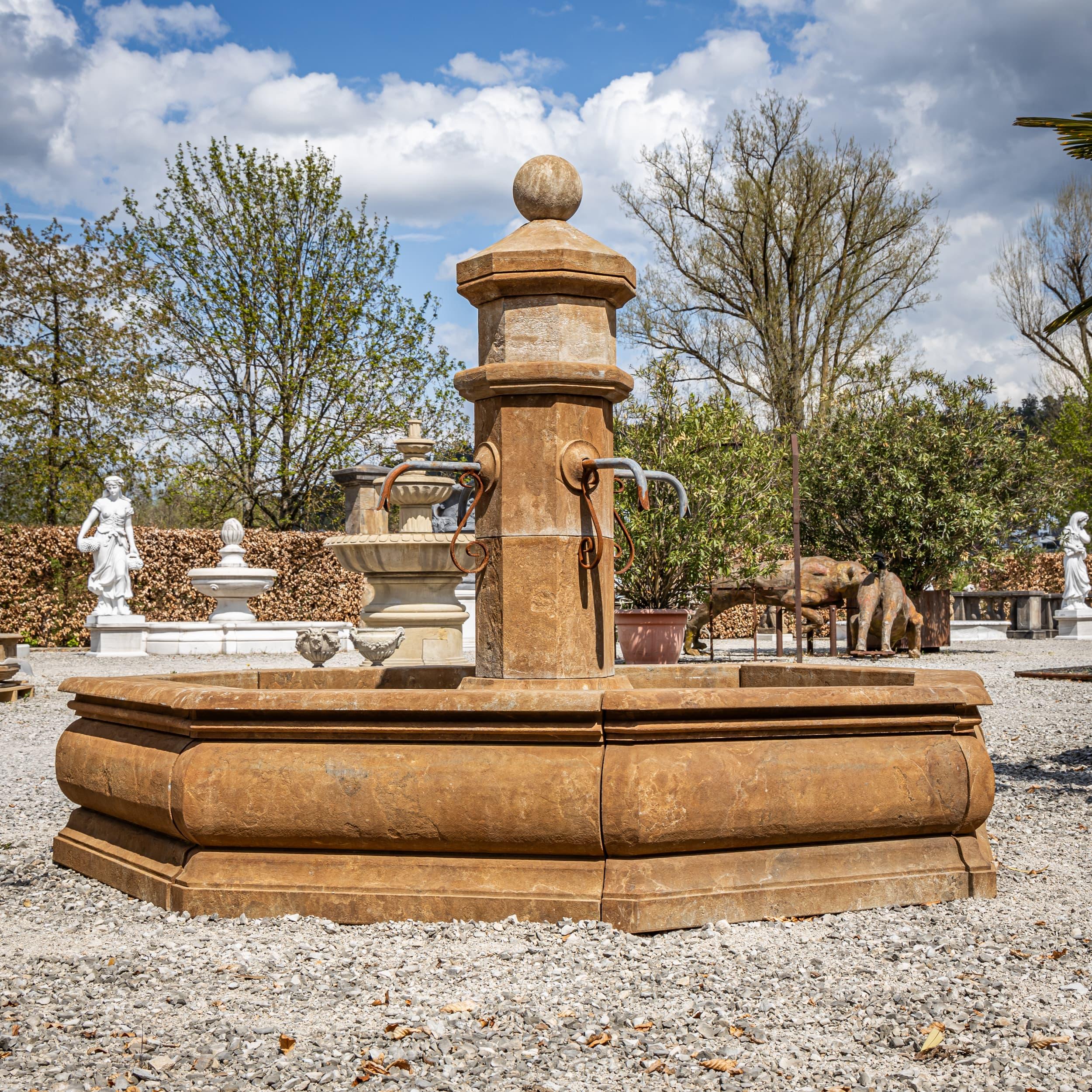 Fuente octogonal de piedra natural cincelada a mano y patinada a la antigua. La columna central está provista de cuatro caños de agua metálicos.