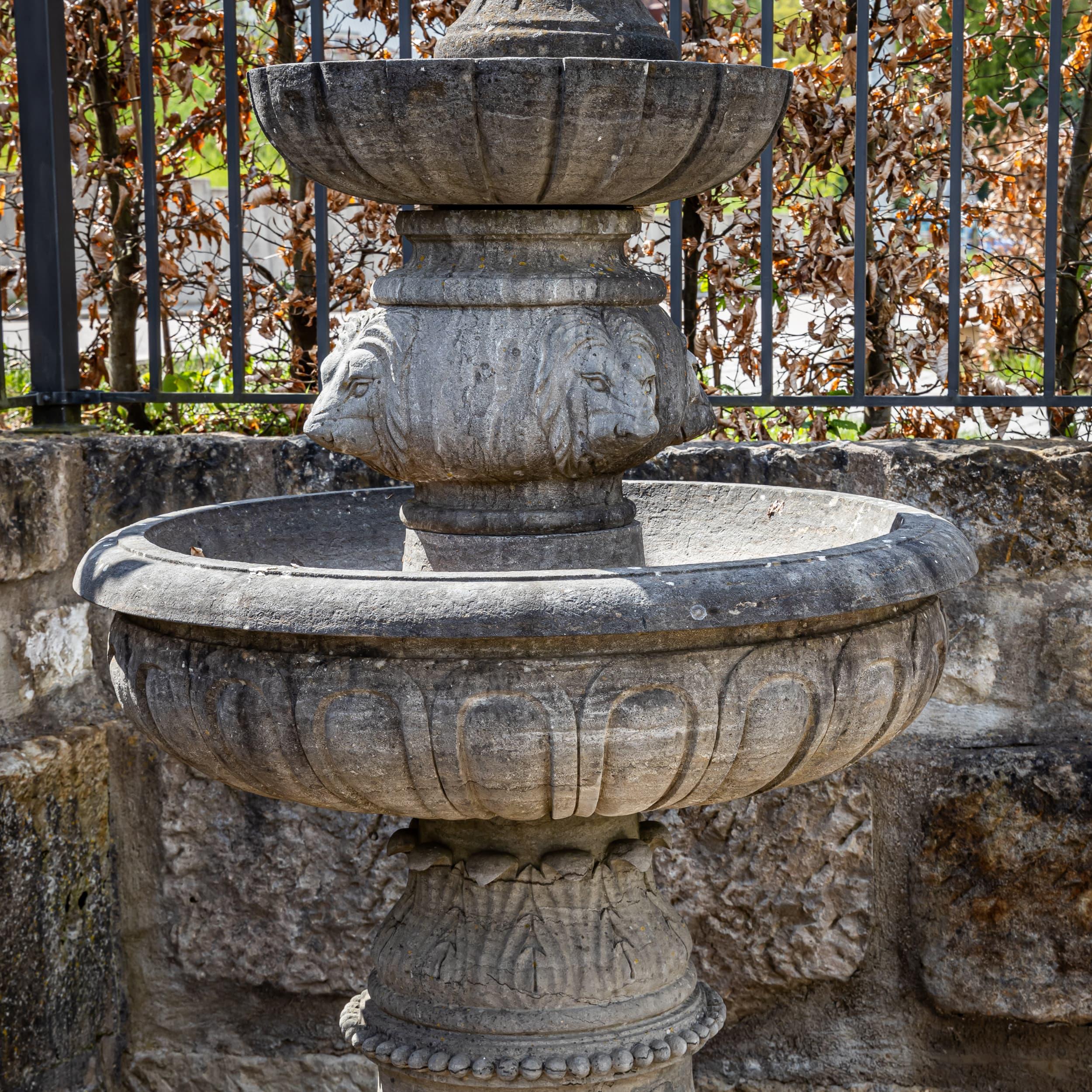 Fountain made of hand-chiseled, antique-patinated natural stone with two stacked bowls. The central section is decorated all around with lion head motifs.