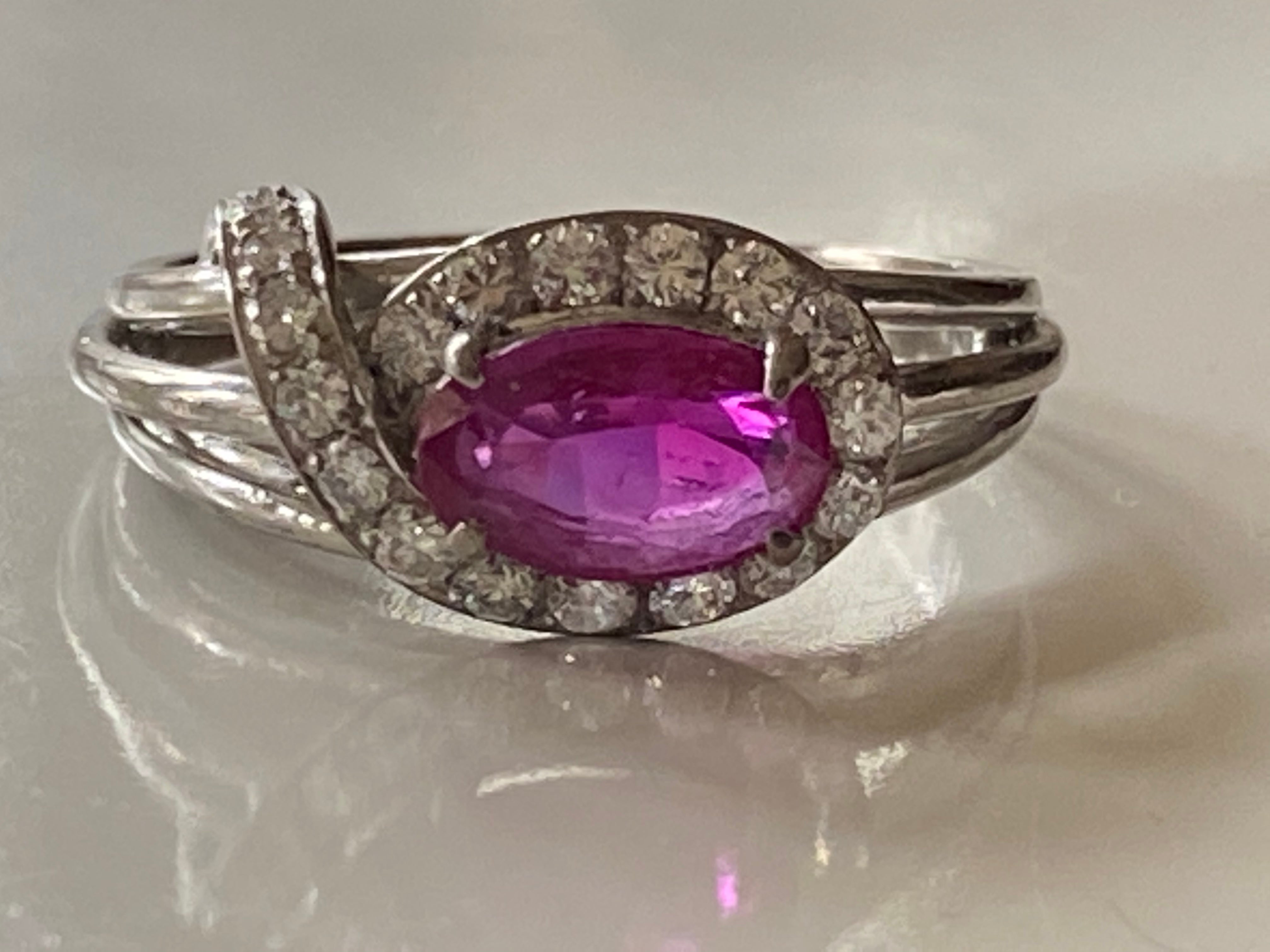 This distinctive ring crafted in the 1960s in platinum is designed around a natural unheated oval-shaped ruby measuring 5.45 x 9mm surrounded by a halo swirl adorned with eighteen gleaming round diamonds totaling approximately 0.50 carats and