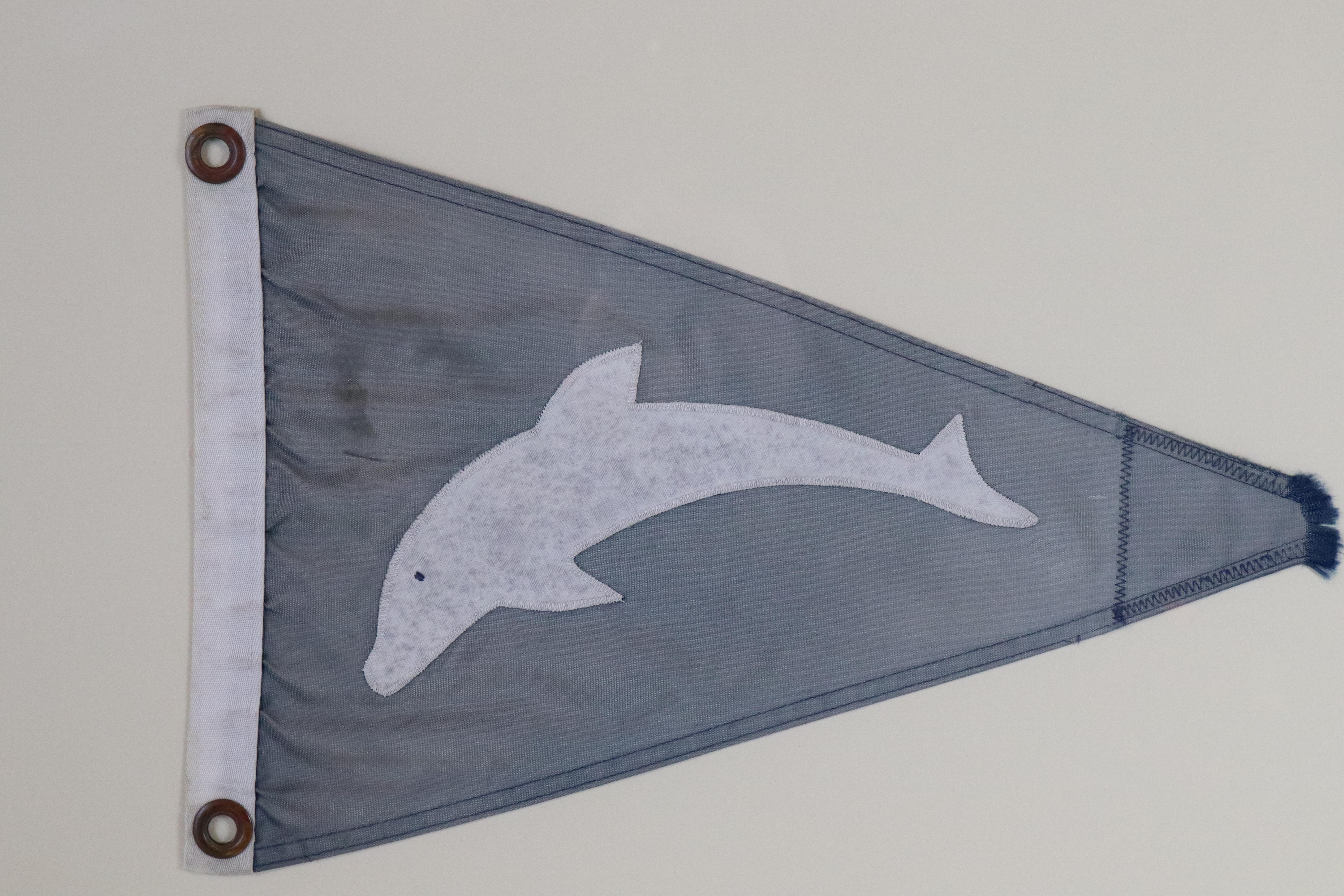 Framed flag showing a white dolphin on a grey/blue field. White hoist with two brass grommets. Matted and framed. Measures: 24