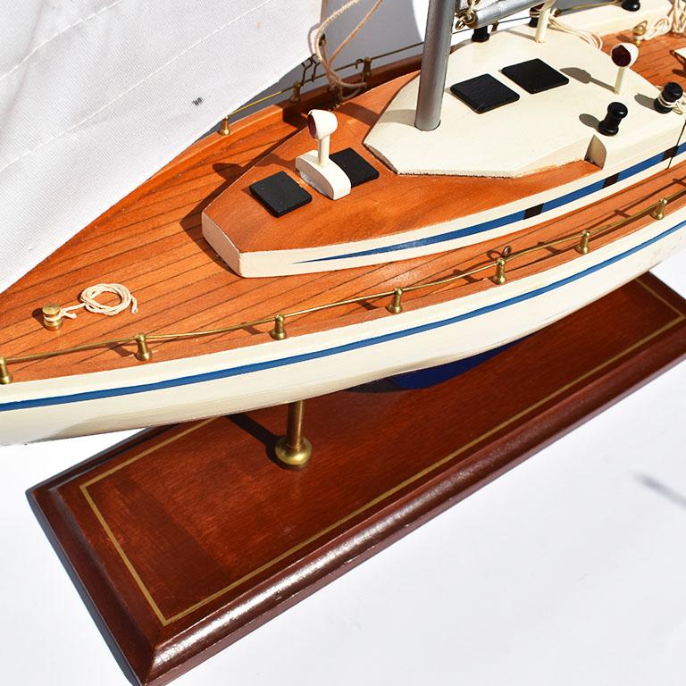 Beautiful model sailboat or yacht created from wood, brass, and canvas. The body is painted in a deep blue at the bottom with white edges. A brass propeller sits underneath. All on a wooden stand. A brass gate with rope surrounds the edges. Two